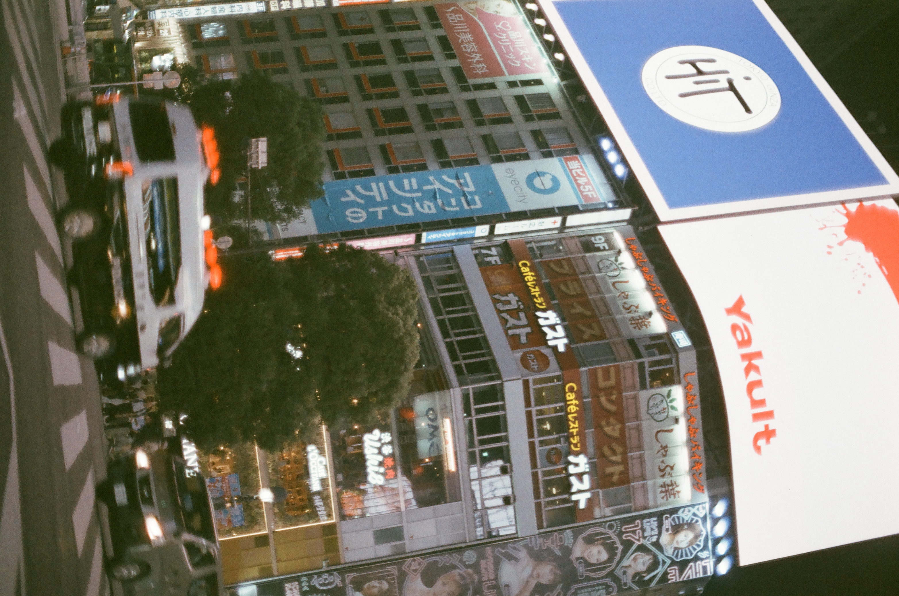 Large billboards reading 'Yakult' and 'HIT' top a tower of restaurant and shop signage above a tree and a passing van.