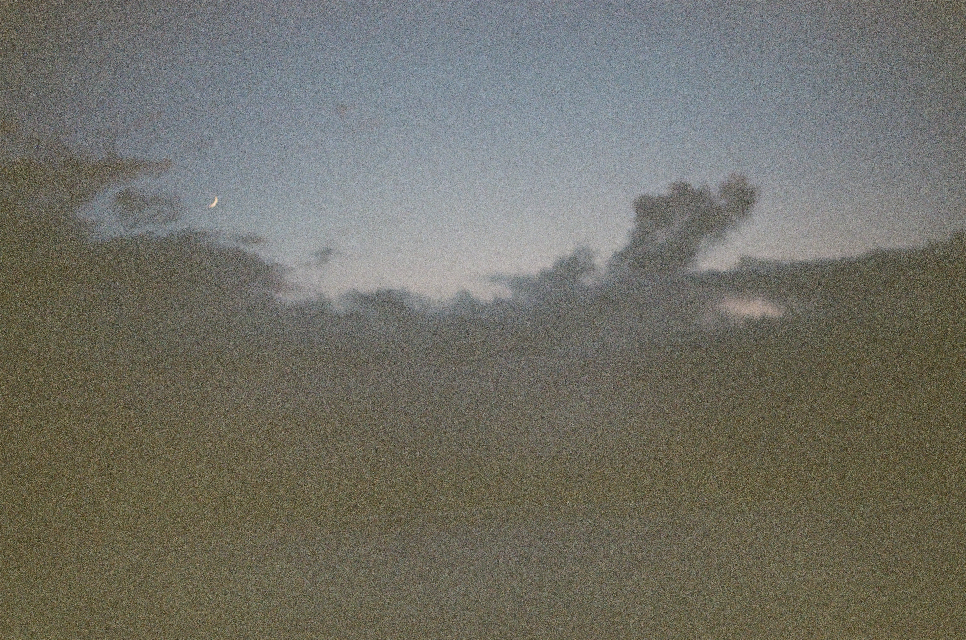A thin crescent moon appears beside dark clouds above layered island forms and a broad dim sea.