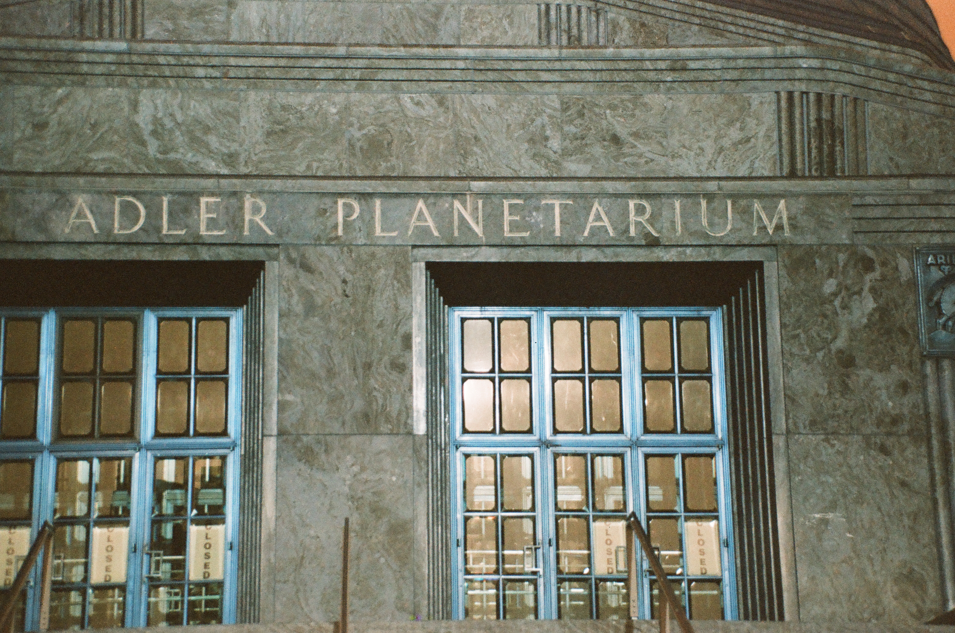 Carved letters spell Adler Planetarium above two sets of blue-framed doors, their glass reflecting light beside handwritten Closed signs.