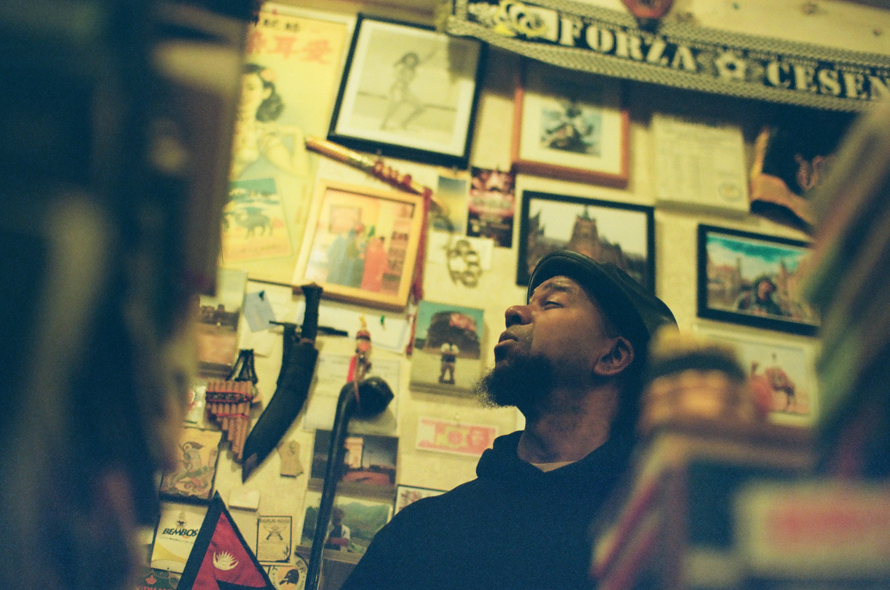 A man in profile looks upward among framed pictures, stacked books, and softly blurred foreground objects.