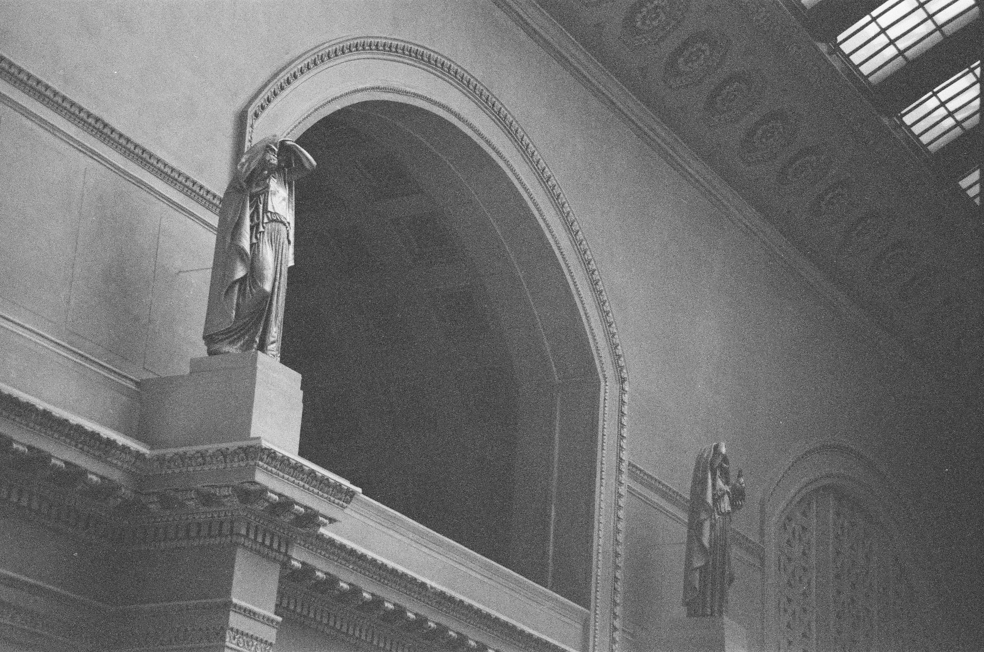 A sculpted figure and layered arch rise toward angled skylights along an ornate monochrome interior wall.