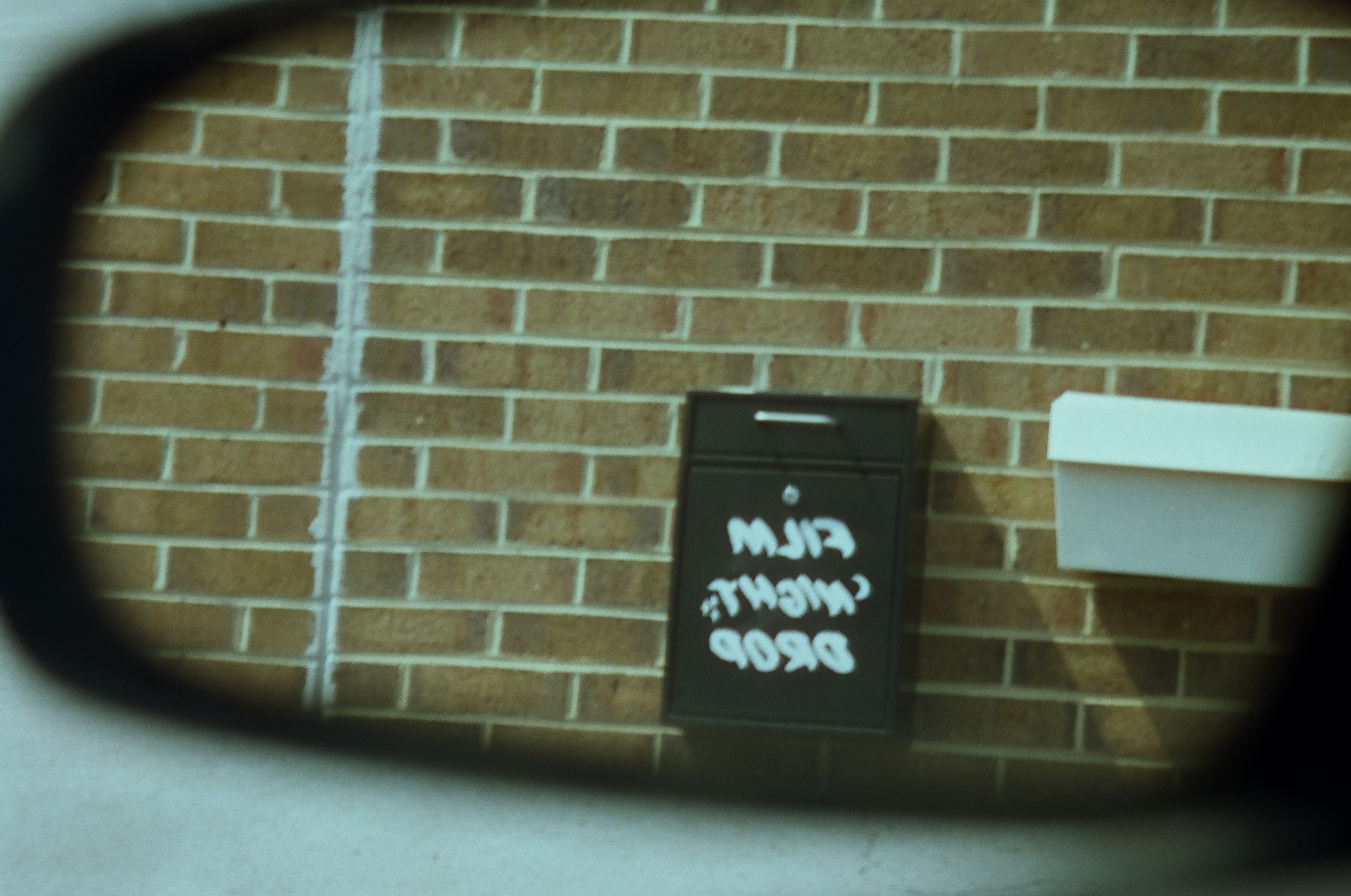 A small black sign with reversed white writing appears in a mirror against a plain brick wall.
