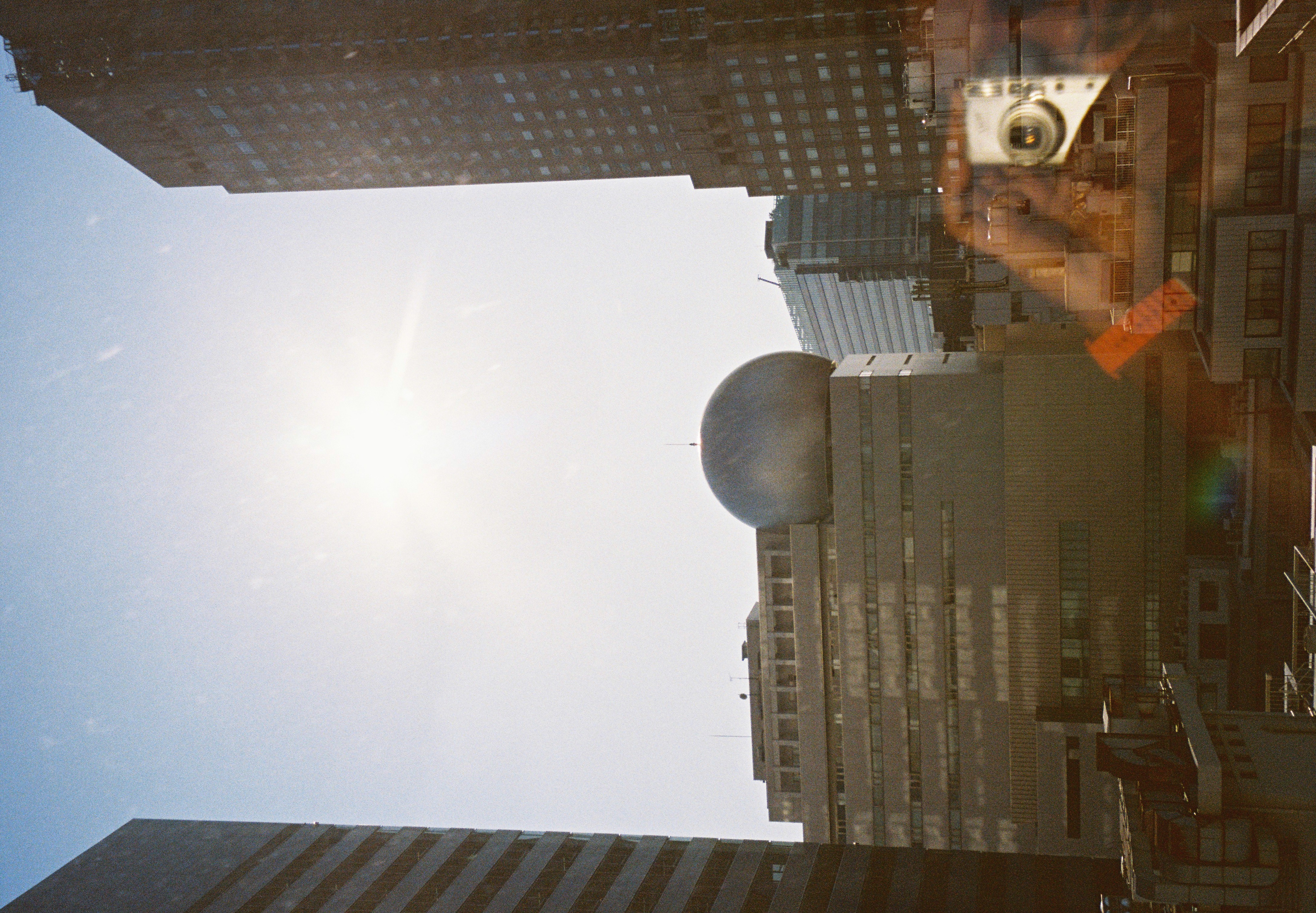 A large silver spherical structure tops a mid-rise building between two dark towers, with a hand and camera reflected in the glass.