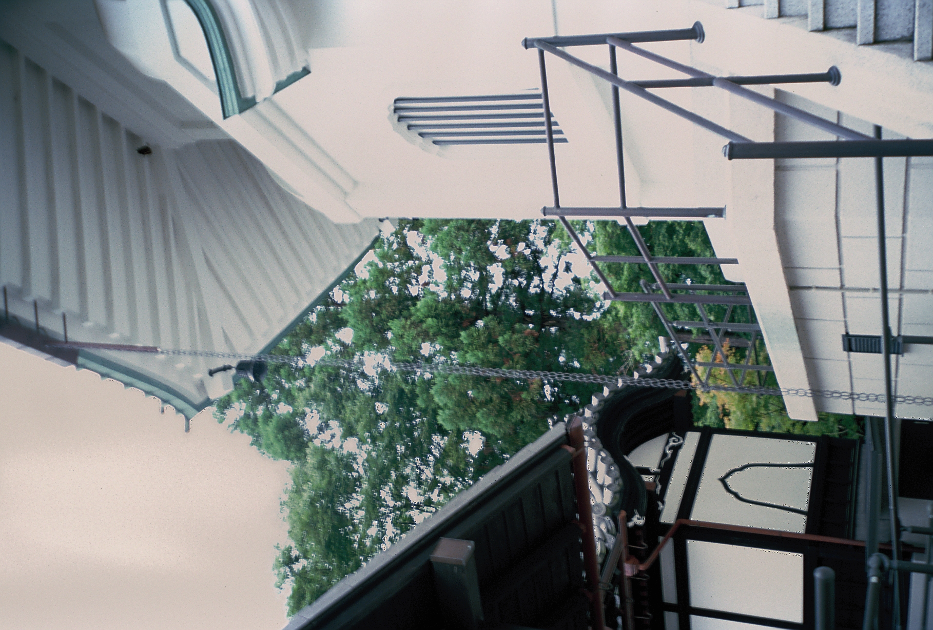 A metal chain hangs from a curved roof eave beside a pale plastered building with a metal stair railing and dense trees behind.