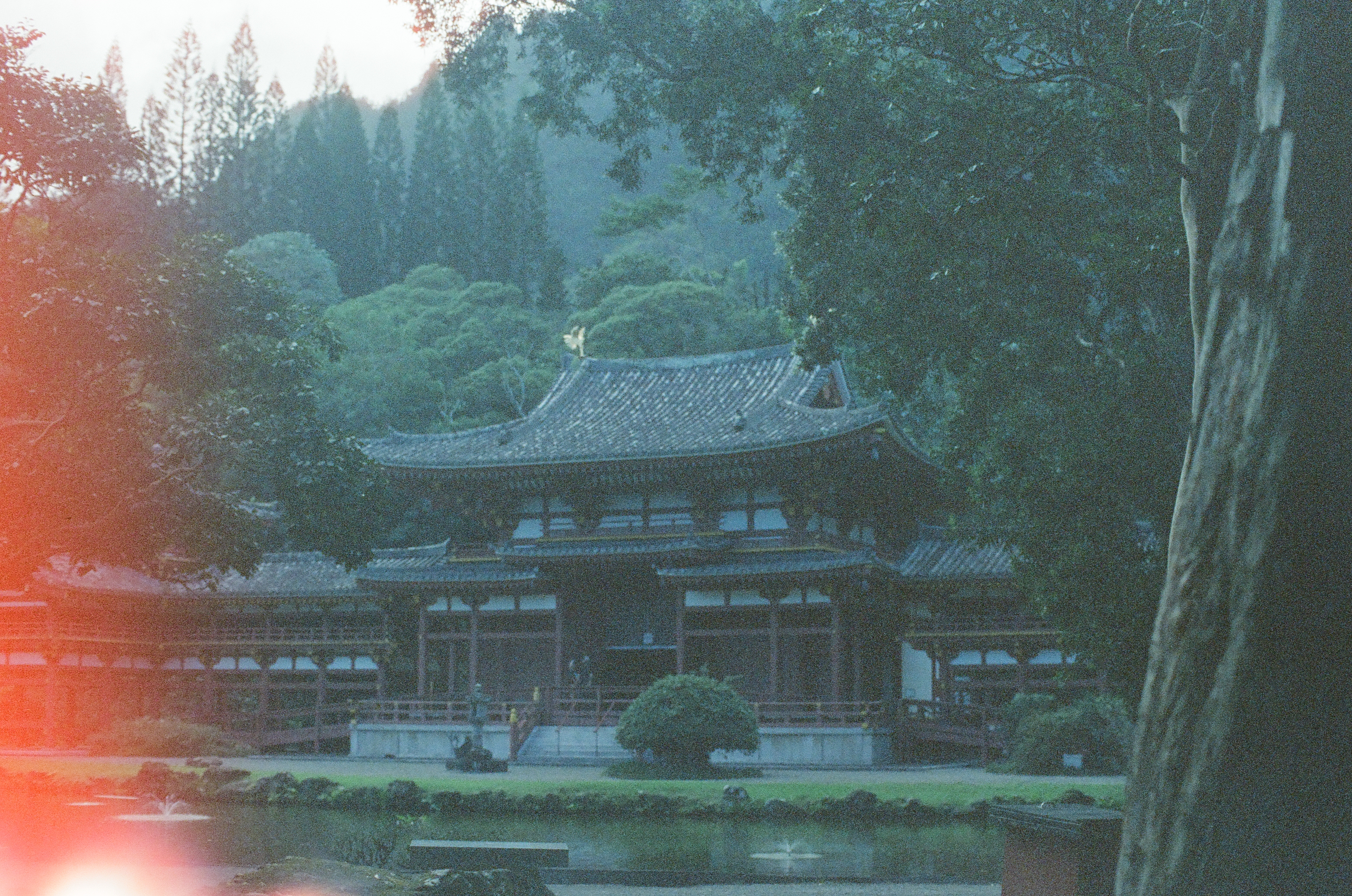 A dark-roofed temple with a gold phoenix finial faces a still pond, backed by misty green hills, with only a small red flare at one corner.