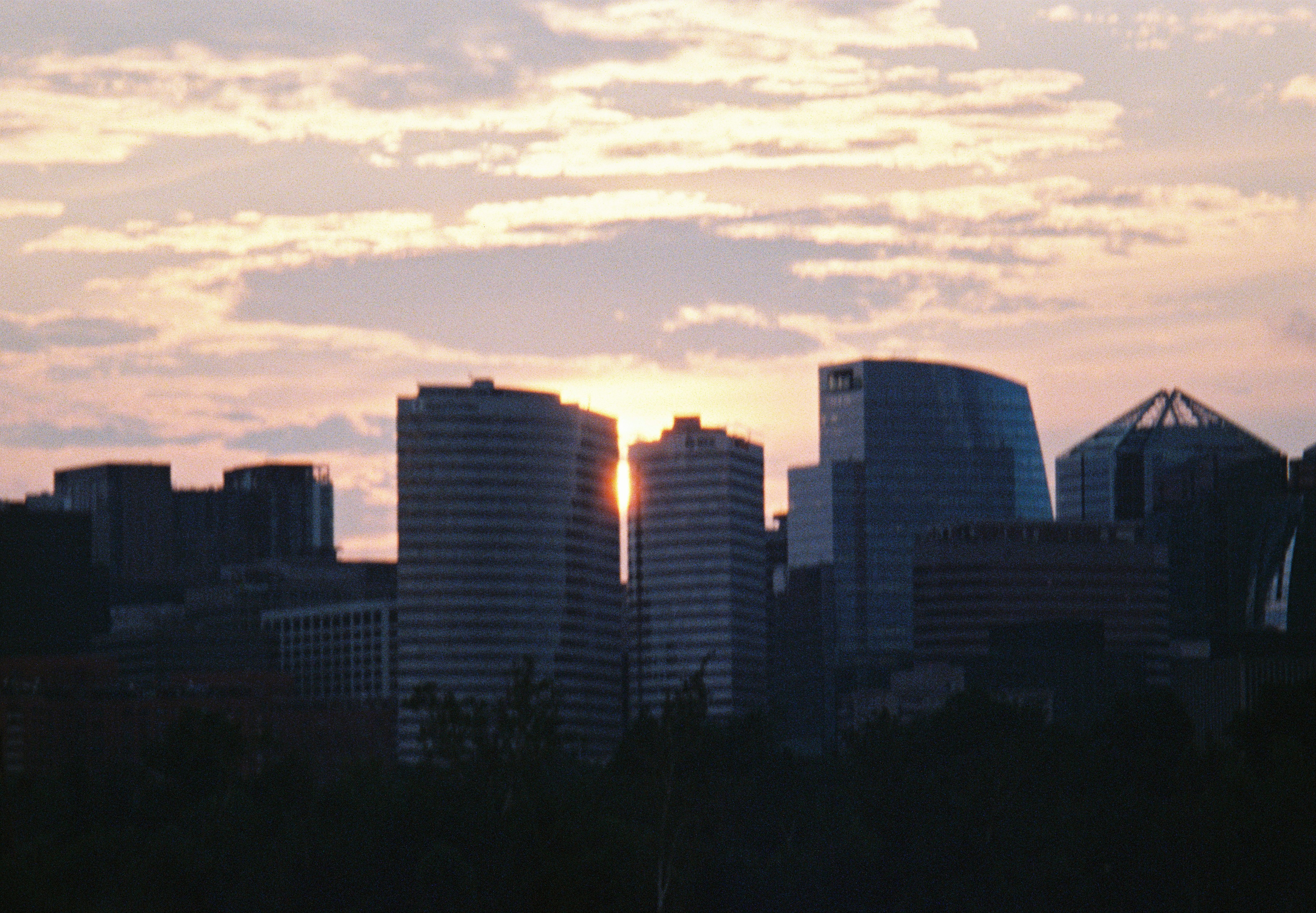 Low sunlight glows between dark office towers beneath a wide peach and blue evening sky.