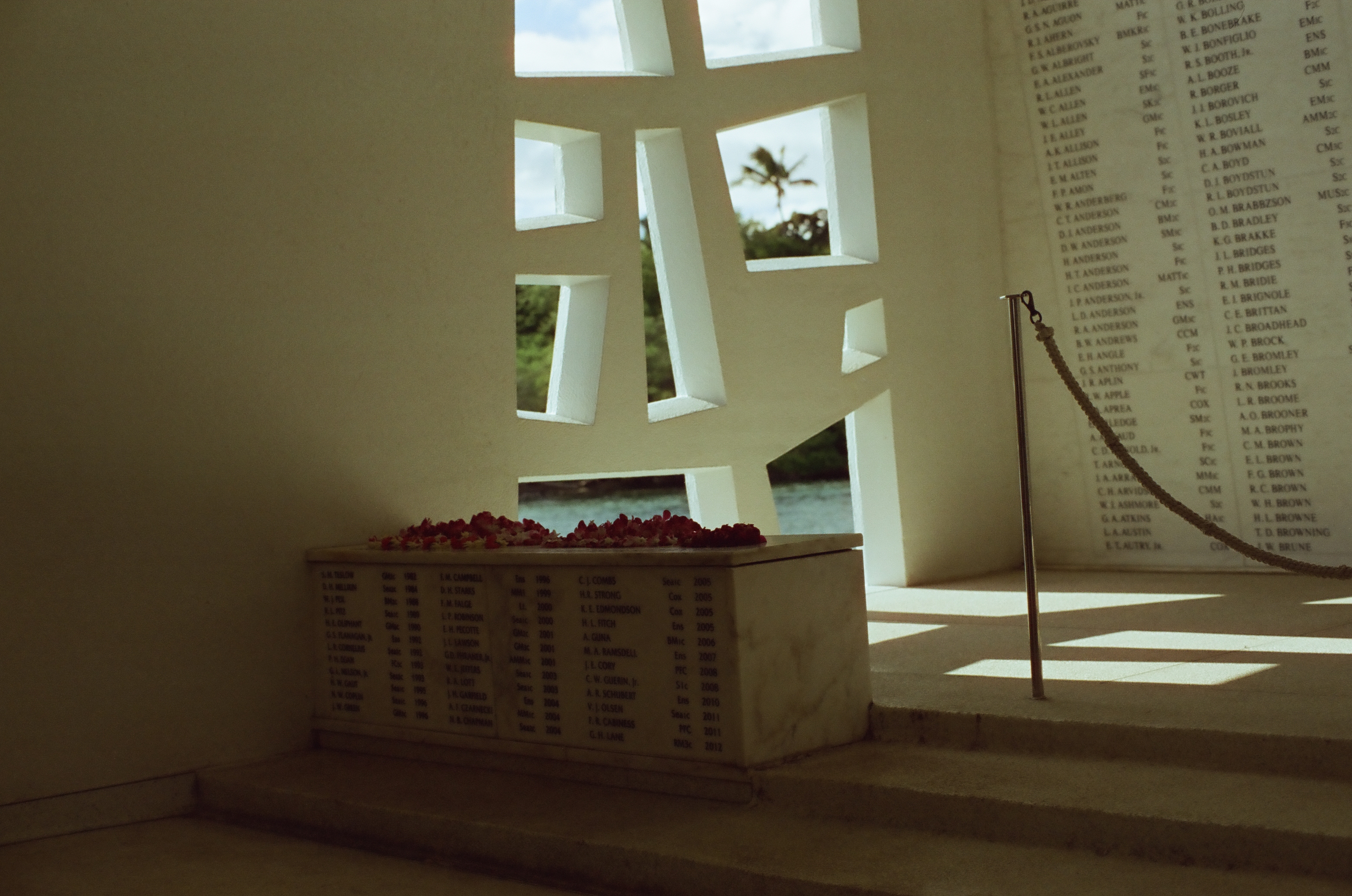 Inscribed names cover pale memorial walls around a flower-topped stone block, rope barrier, and geometric windows.
