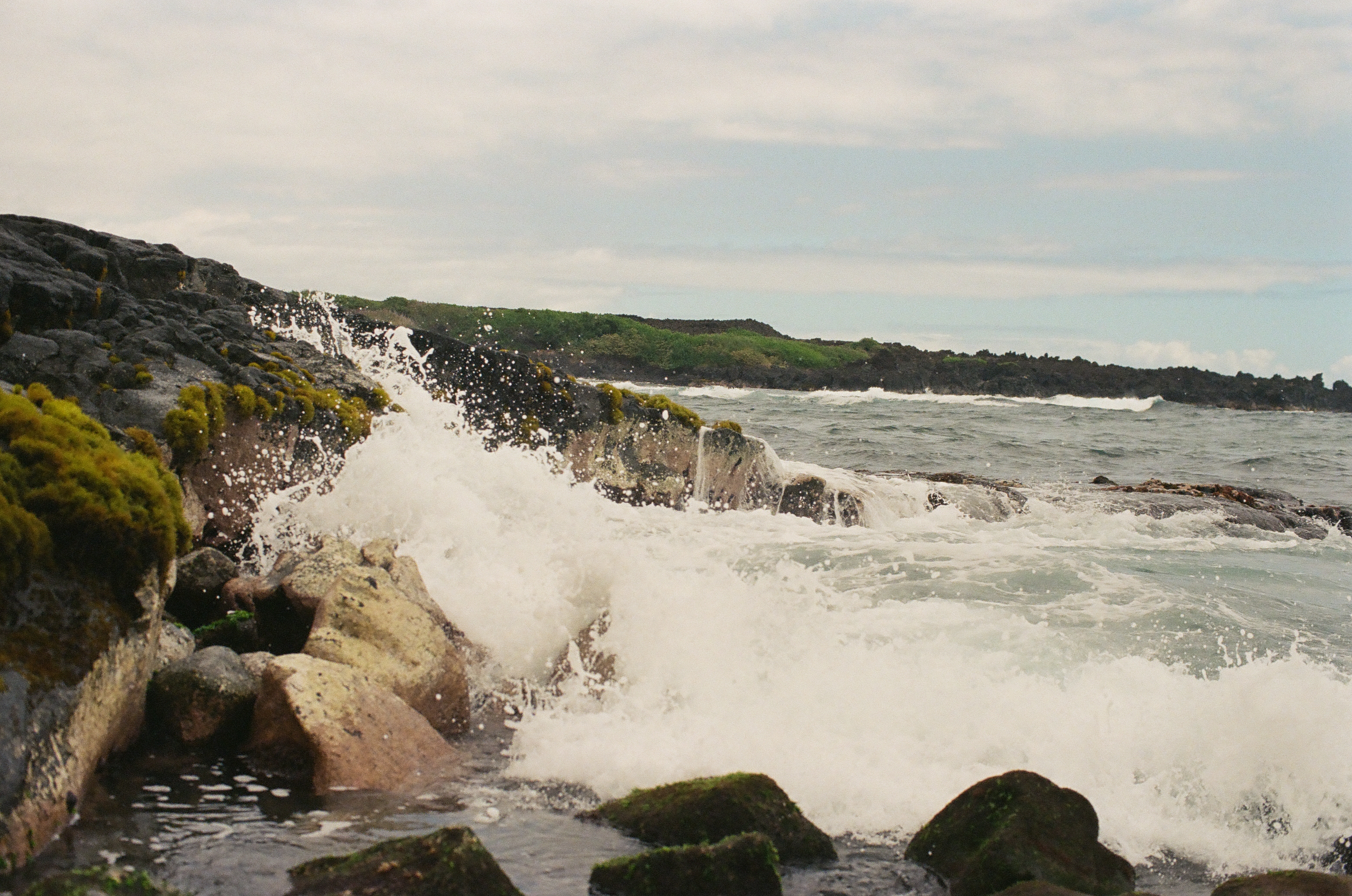 Whitewater surges up and over black rocks thick with yellow-green moss, spraying across a low headland toward the open sea beyond.