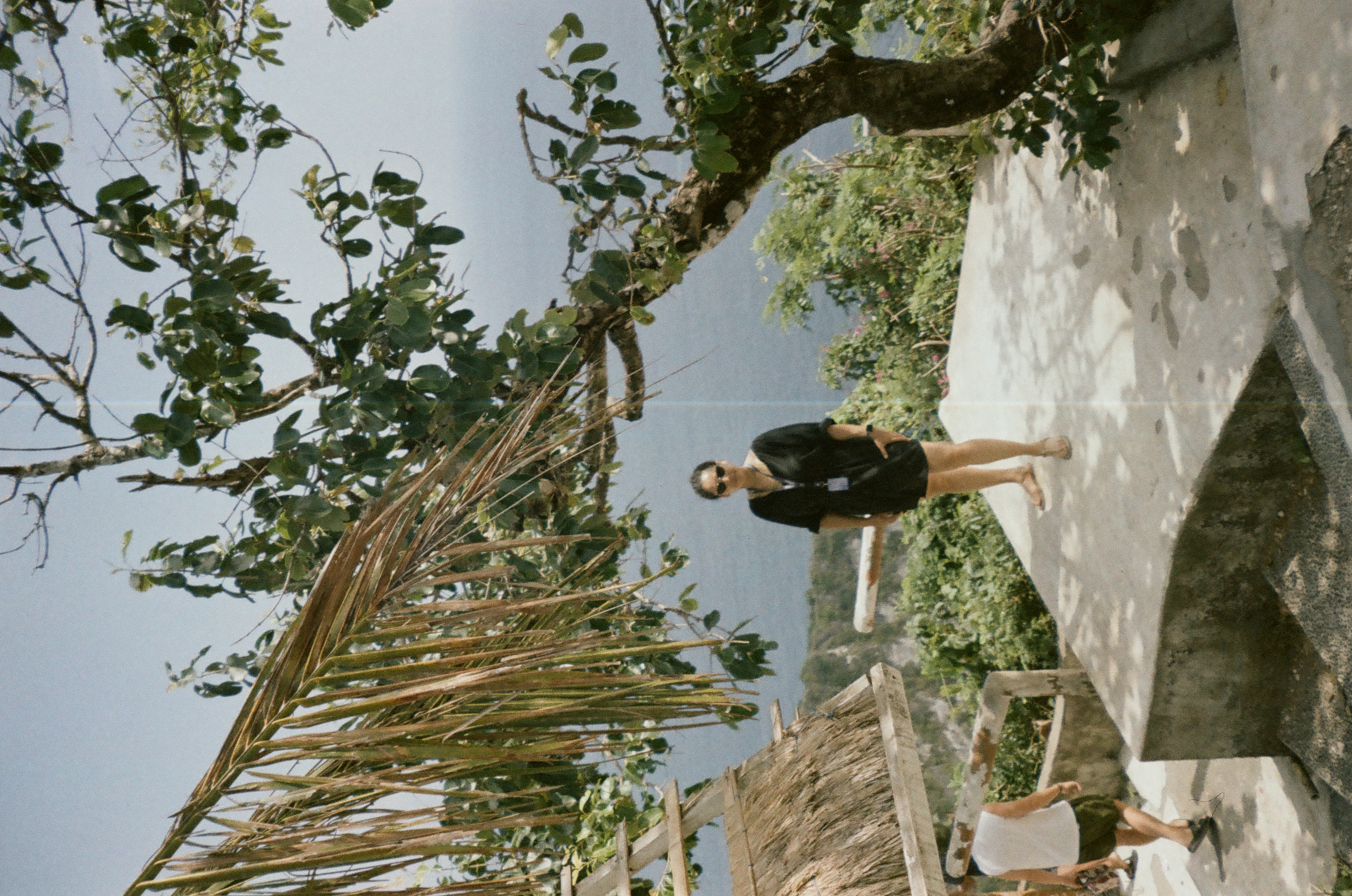 A person stands on a bright terrace among leafy branches, a thatched roof, and distant water.