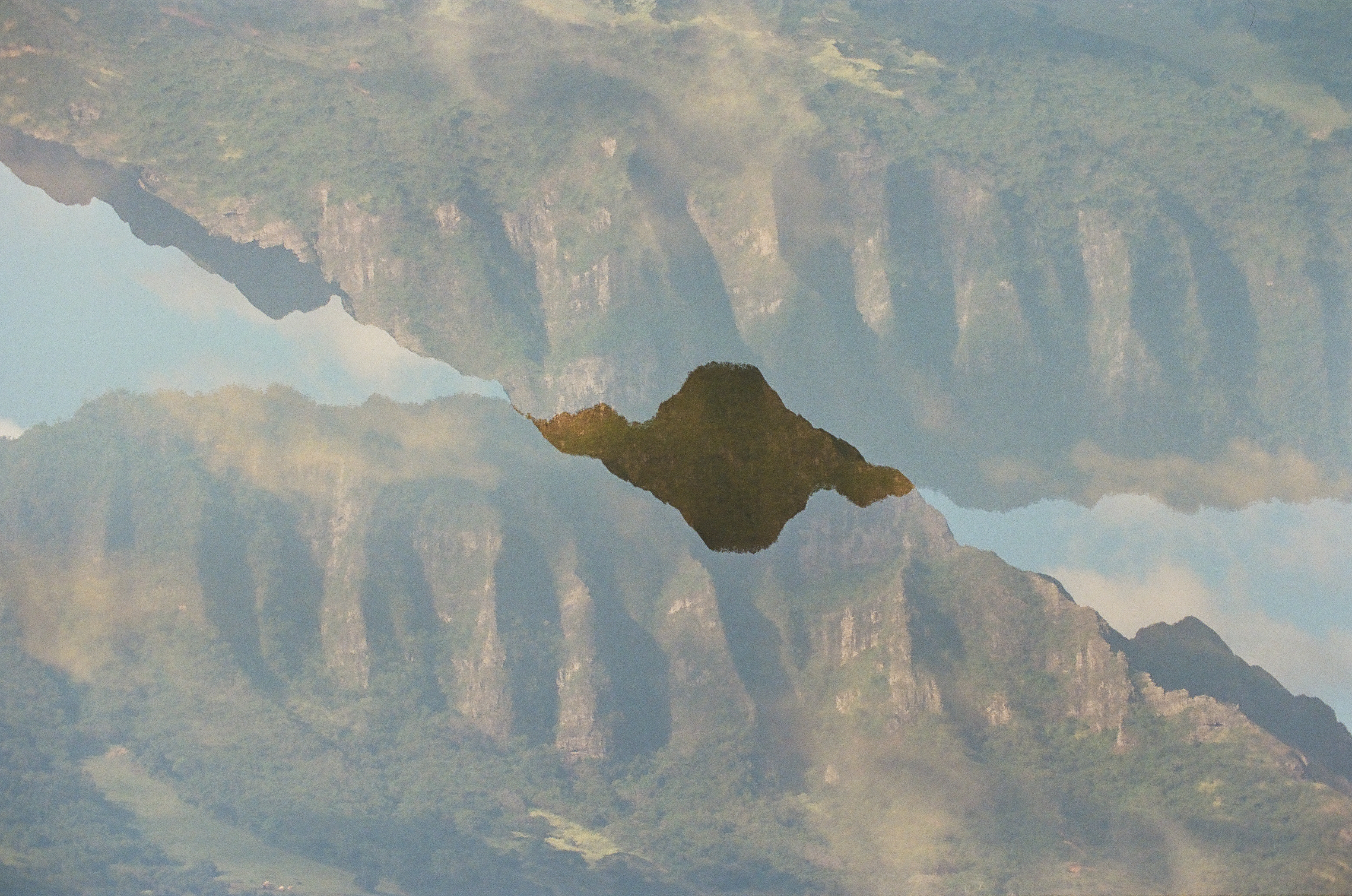A double exposure mirrors the ridged green mountains and a dark island shape, one inverted atop the other in a symmetrical reflection.