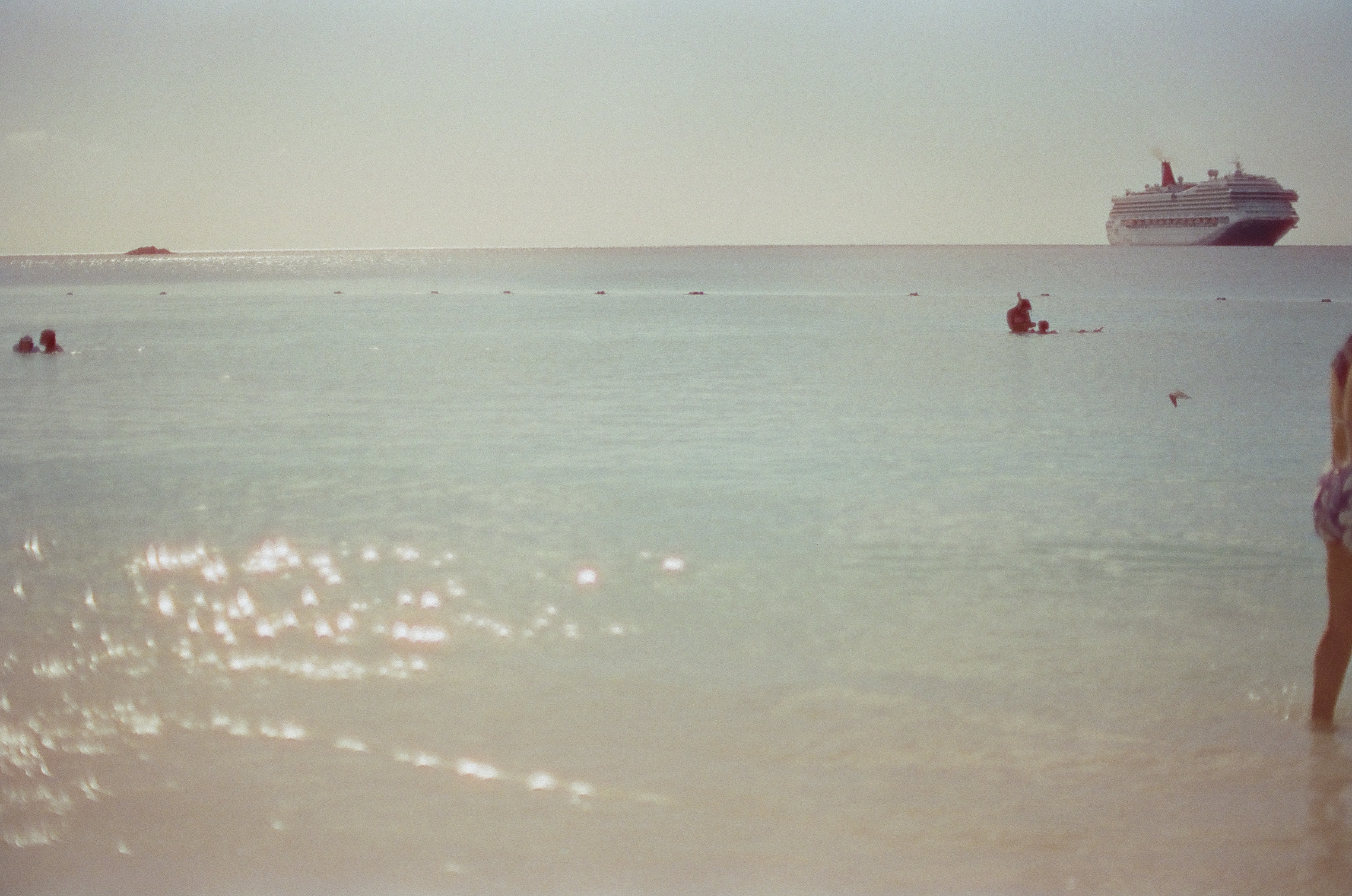 Sunlit shallow water extends toward swimmers, a buoy line, and a large ship, with part of a standing adult cropped at right.