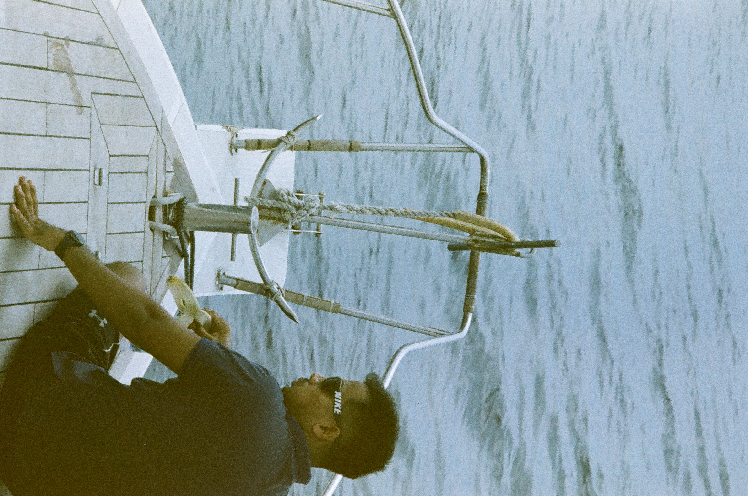 A seated person in dark clothing leans near the bow rail while pale blue water fills the background.