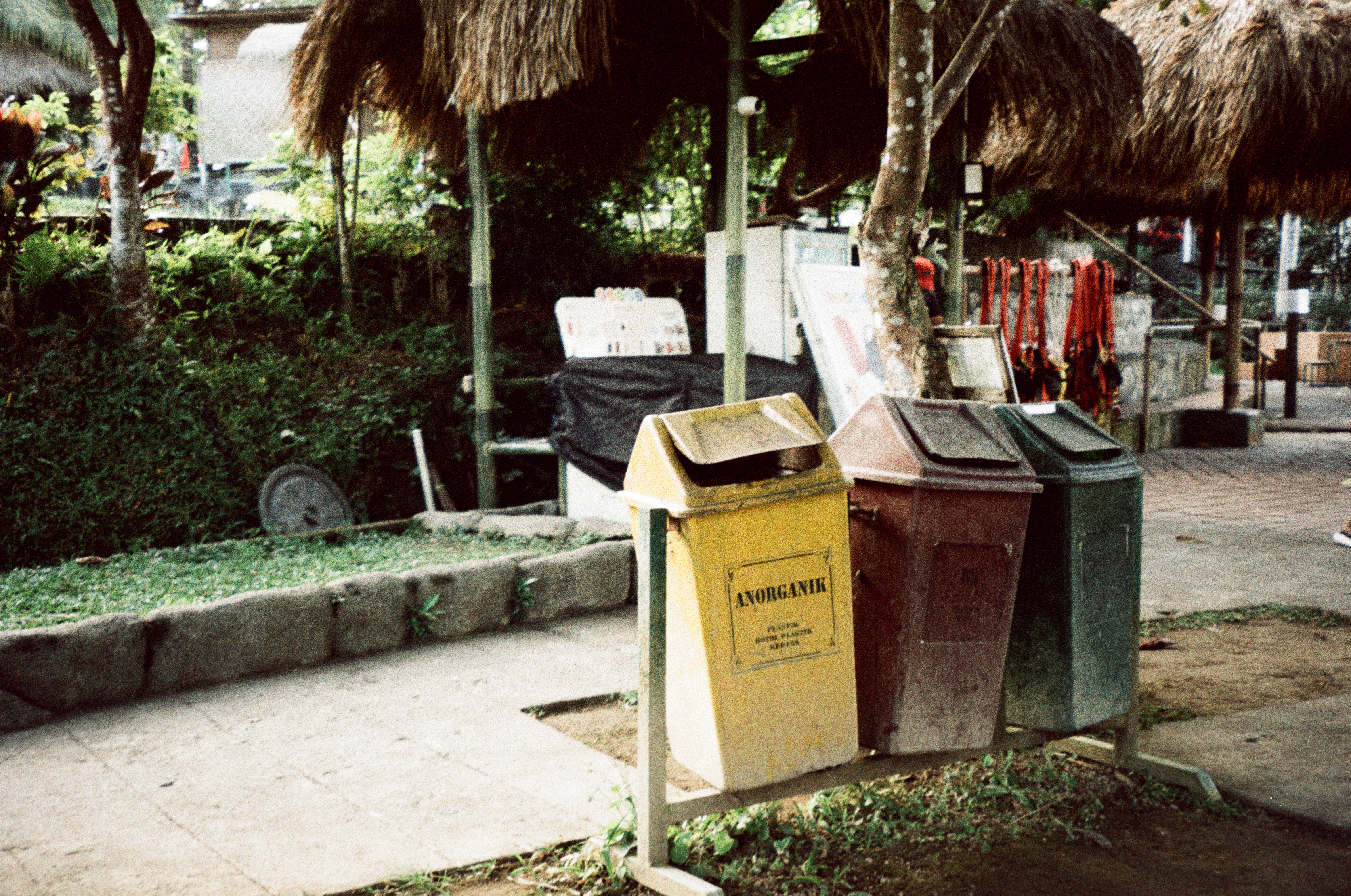 Yellow, red, and green bins stand on a shared rack near thatched shelters, with “ANORGANIK” readable on the yellow bin.