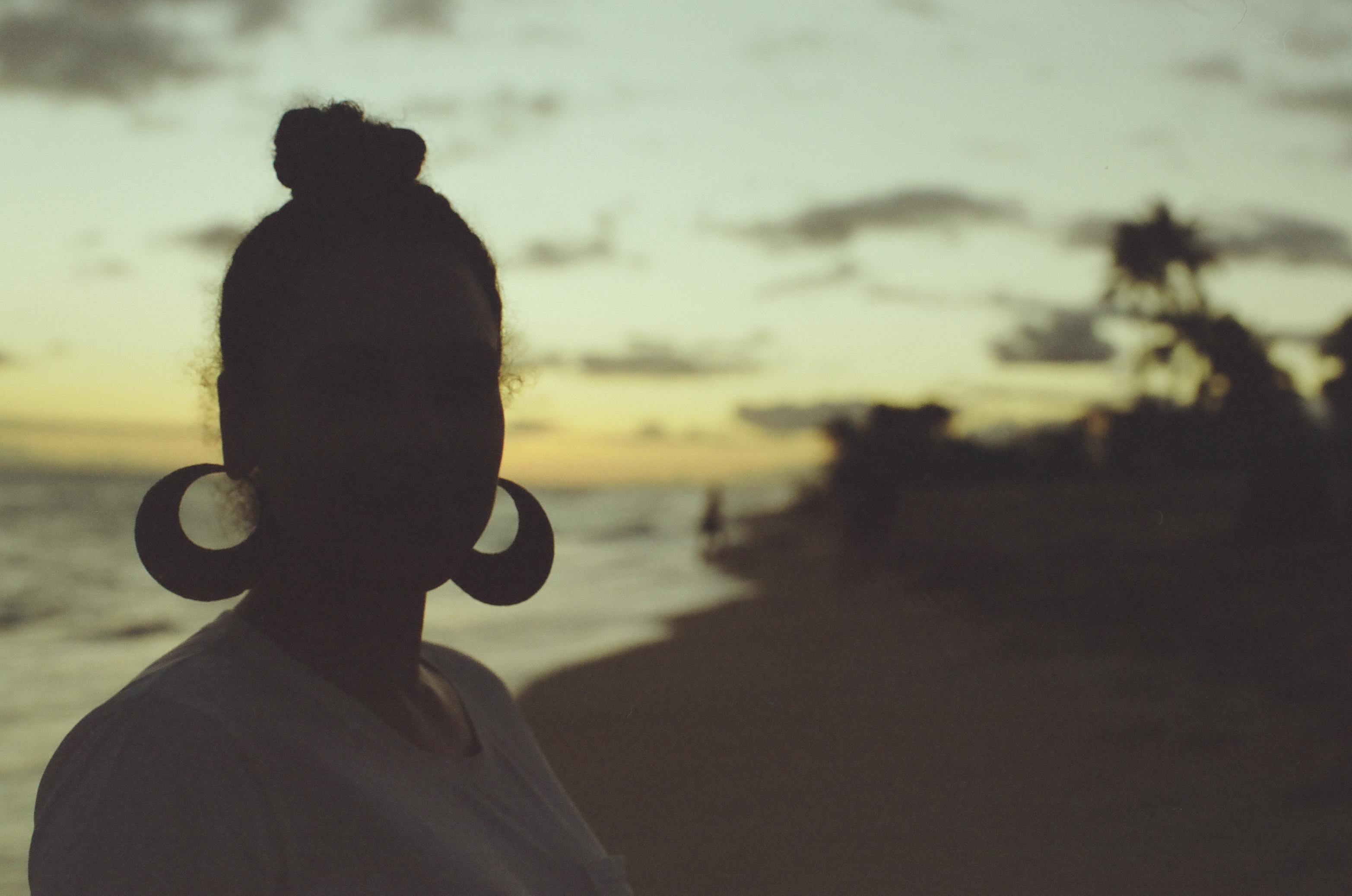A person with hair tied high and large round earrings appears in silhouette against the evening water.