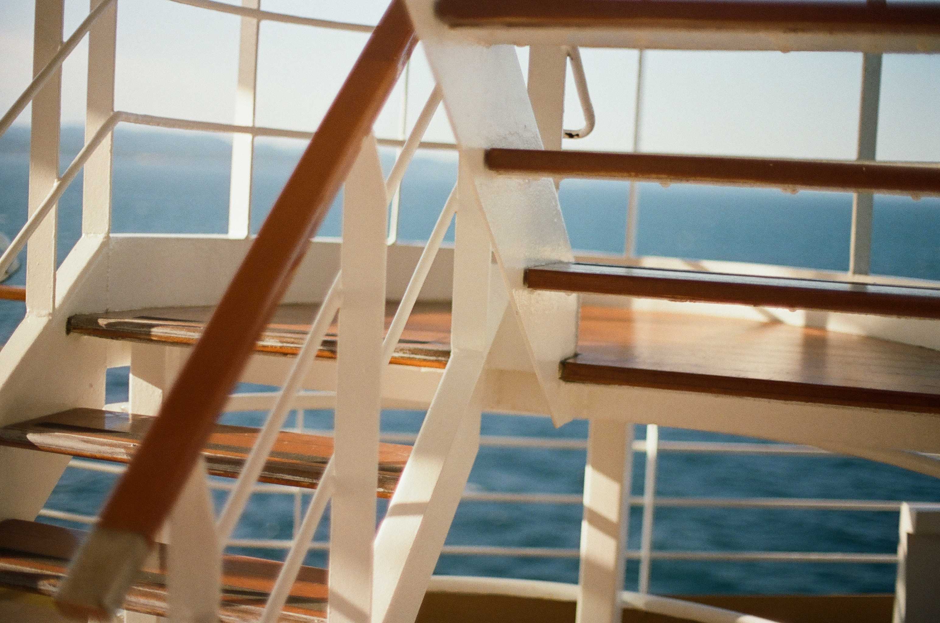 White exterior stairs with dark wood treads and painted metal railings ascend above a blurred view of open blue ocean and distant coastline.