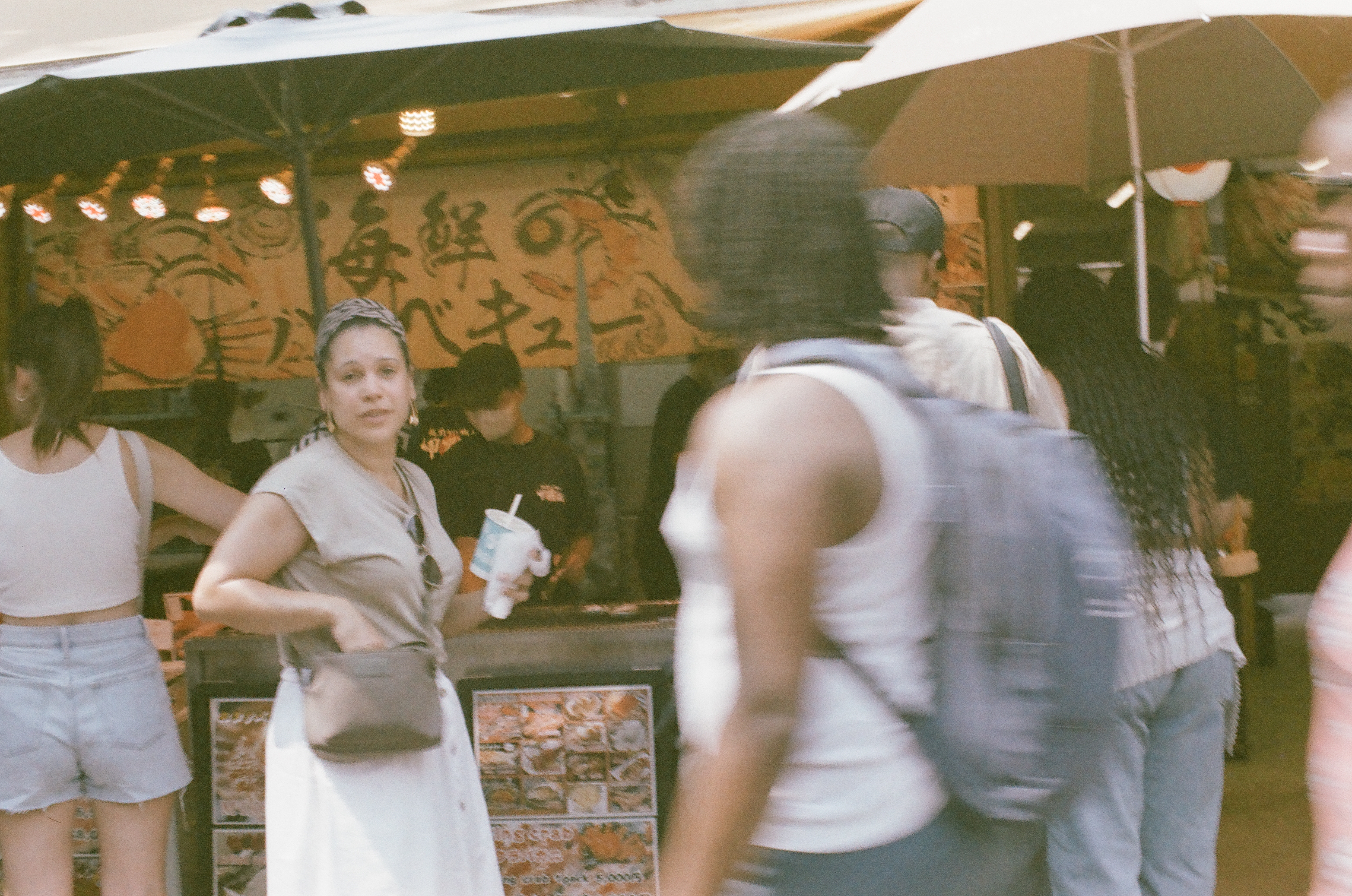 Iced drink in hand, a woman with a patterned headscarf stands beside a hand-painted seafood stall while a blurred figure with a backpack crosses the foreground.