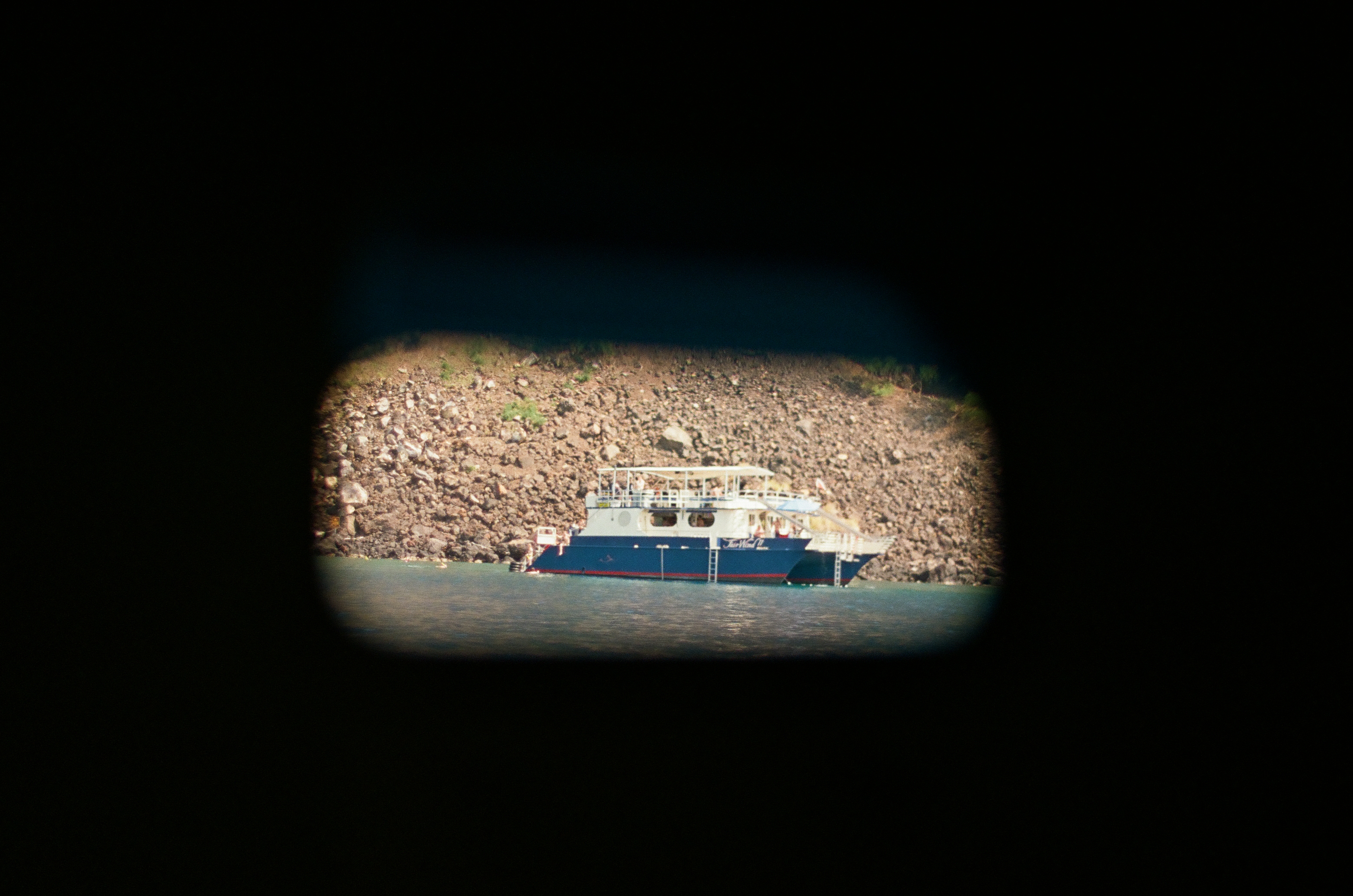 A blue and white boat appears through a small rounded opening surrounded by deep black foreground and rocky shore.