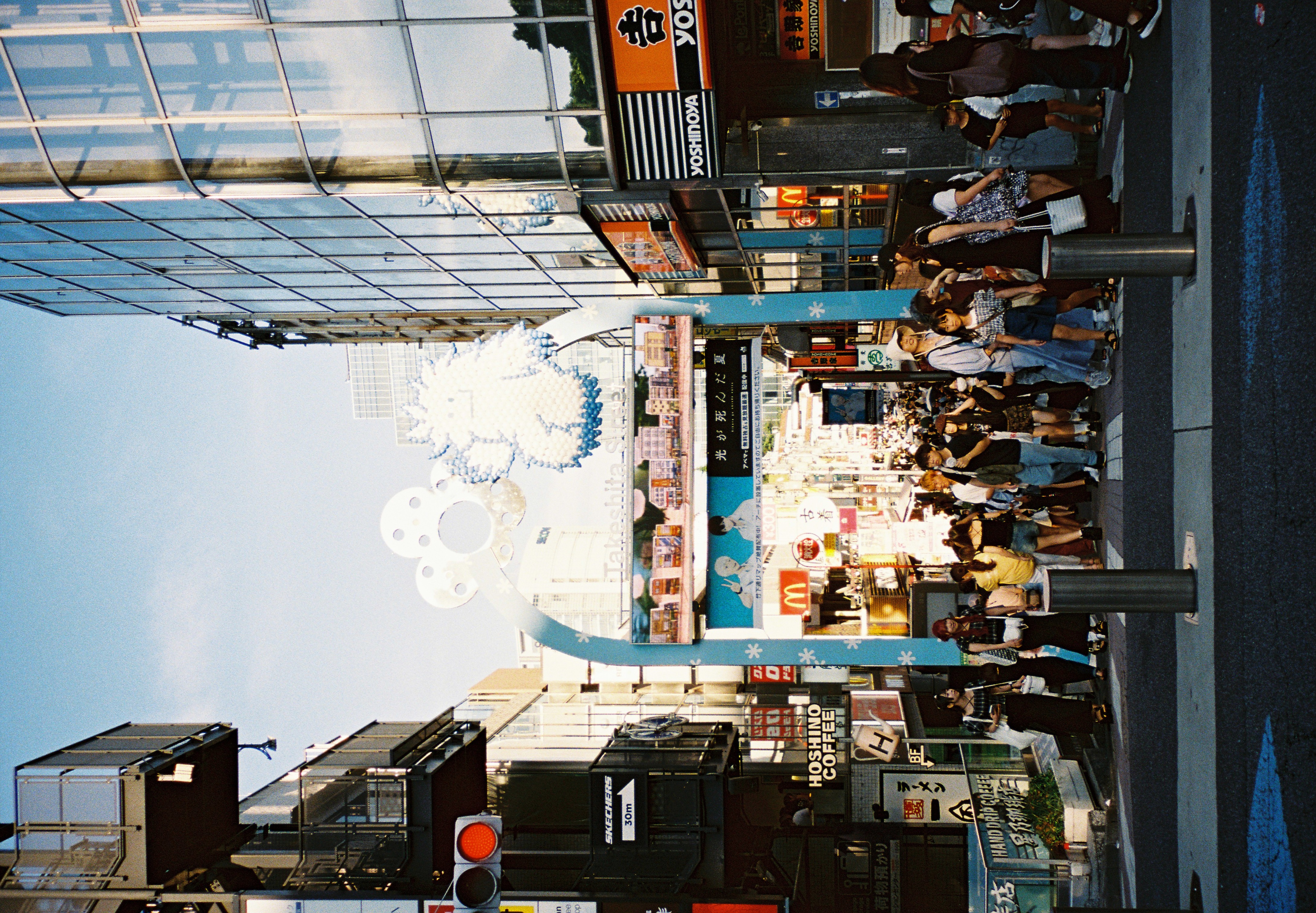 A pale blue arch topped with white sculptural shapes frames a crowded pedestrian street between a glass tower and stacked shop signage.