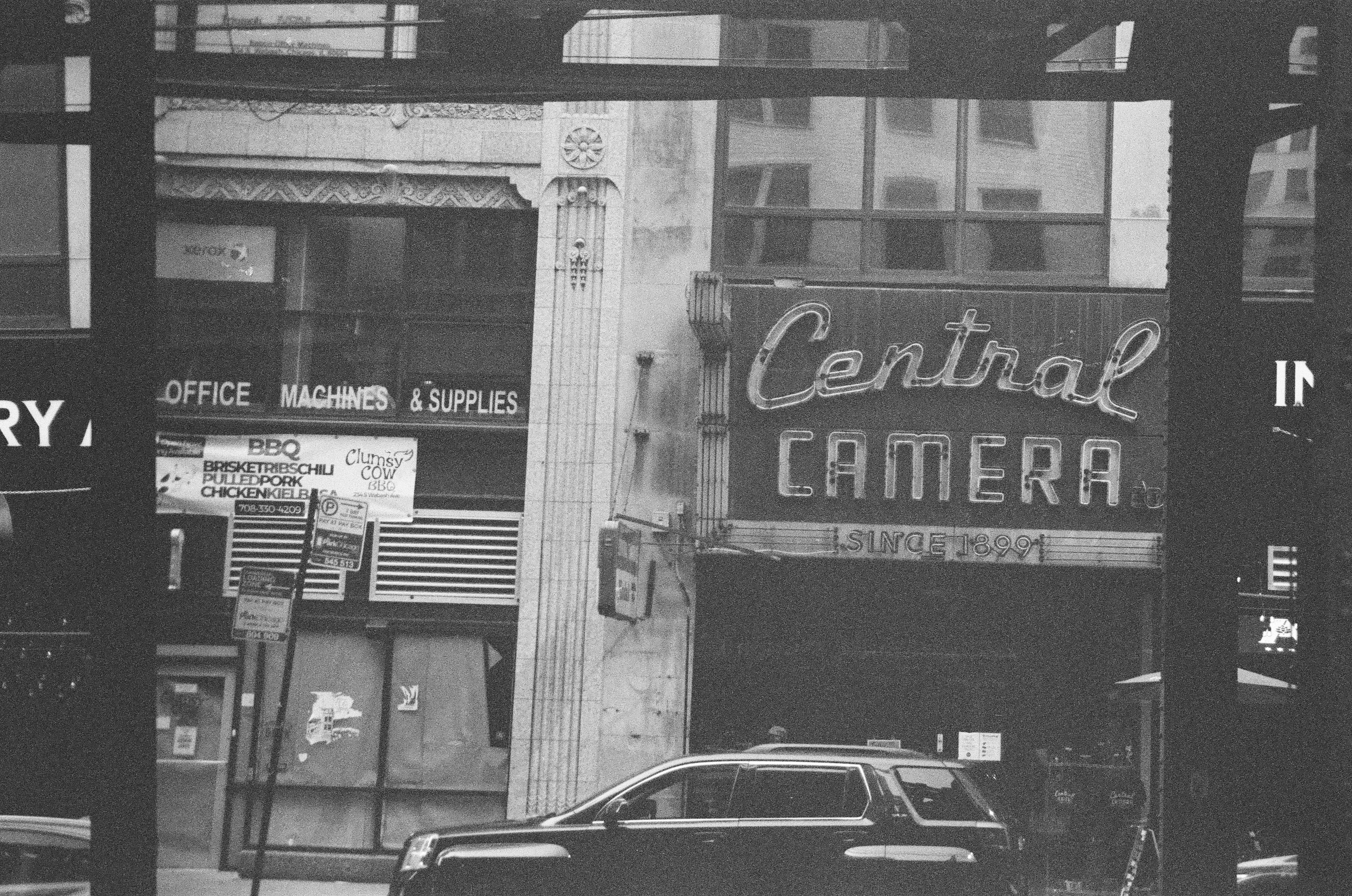 A monochrome storefront with a large camera-shop sign rises behind a dark parked car and shadowed street columns.