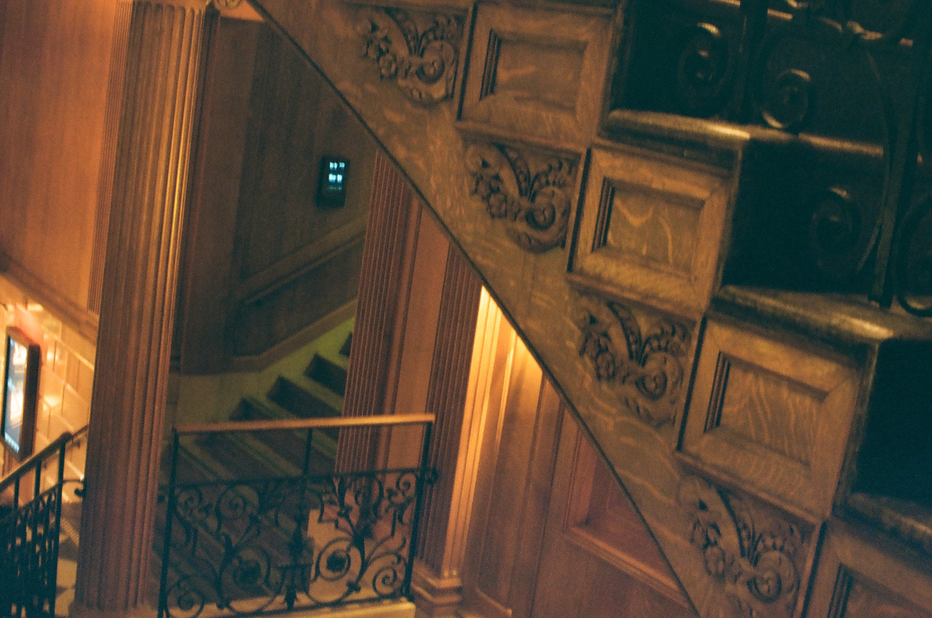 A paneled staircase with an ornate wrought iron railing descends past carved wood columns toward a small illuminated wall panel.