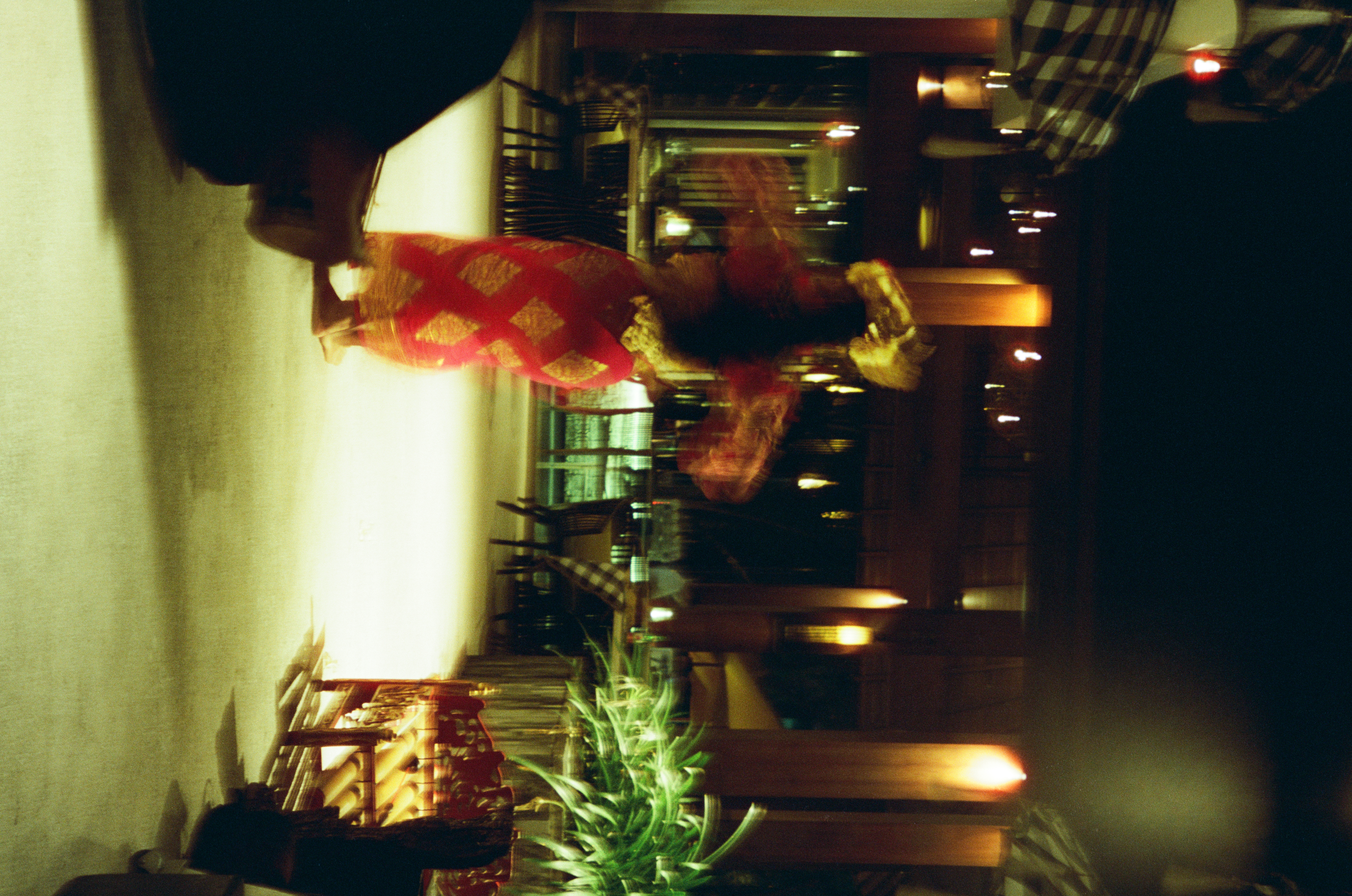 A motion-blurred figure in red and gold with a large gold head adornment raises both arms on a bright floor among dark tables and warm lights.