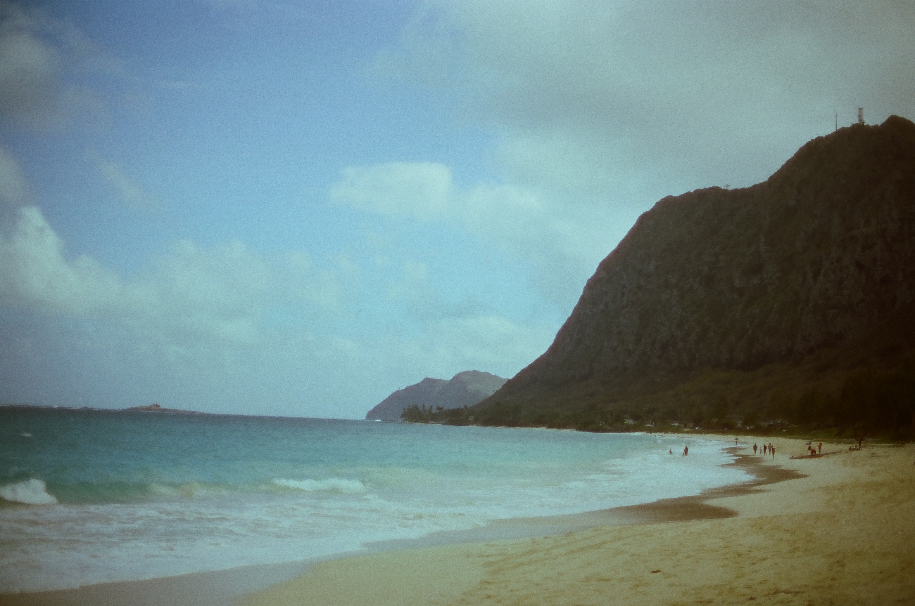 A sandy beach curves toward a steep dark ridge while small swimmers and walkers gather along turquoise water.