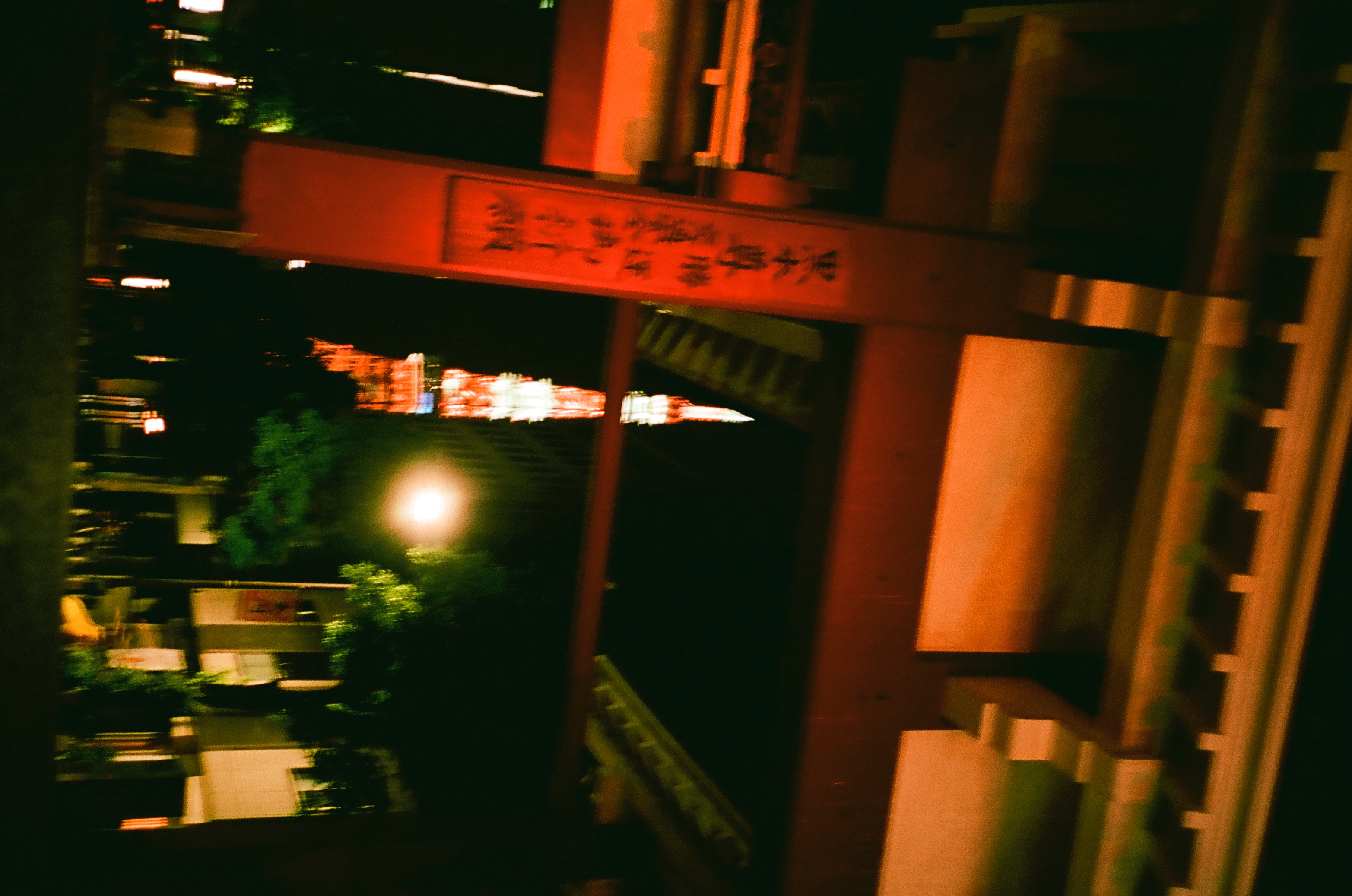 A large red wooden gate structure with an inscribed pillar frames a distant, blurred lattice tower beside a bright halo of lamplight.