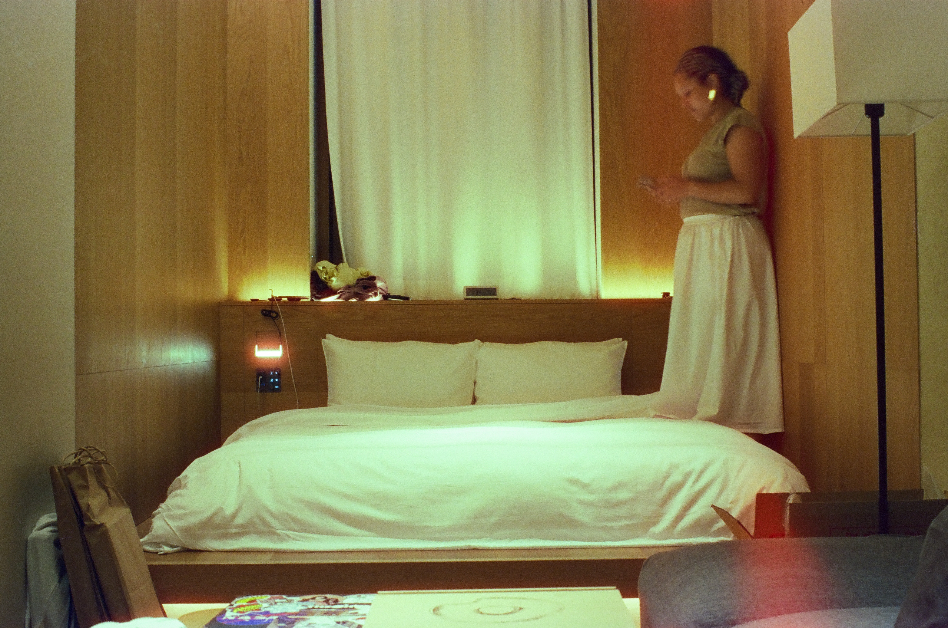 A woman in a sleeveless top and long white skirt stands beside a white bed against a wood-paneled wall, looking down at a small handheld device.