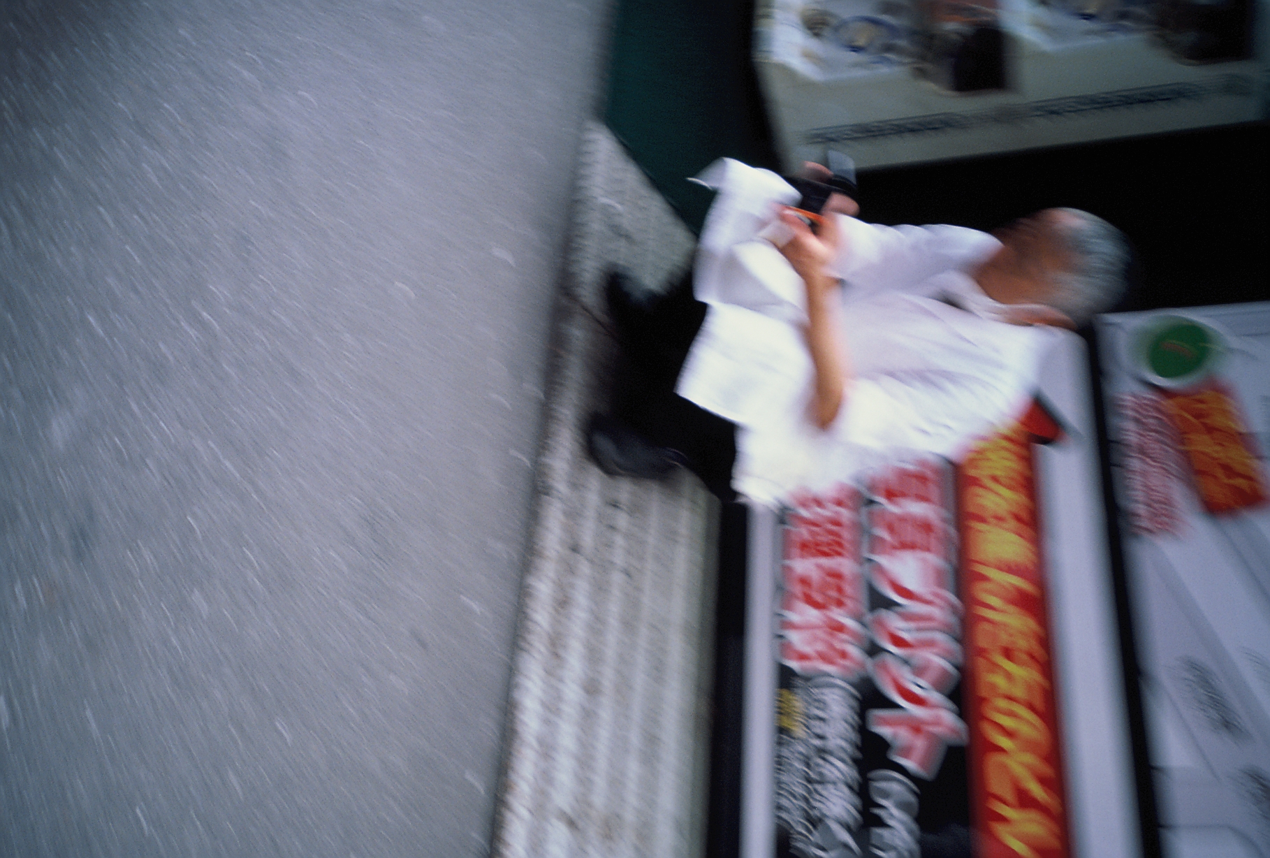 A gray-haired man in a white jacket crouches on a step, his motion blurred beside red signage with white Japanese lettering.