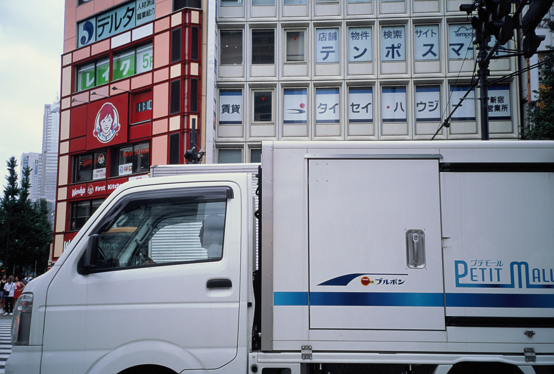 A white box truck advertising a snack brand rolls past a red storefront and a tall building covered in Japanese signage, with wires overhead.