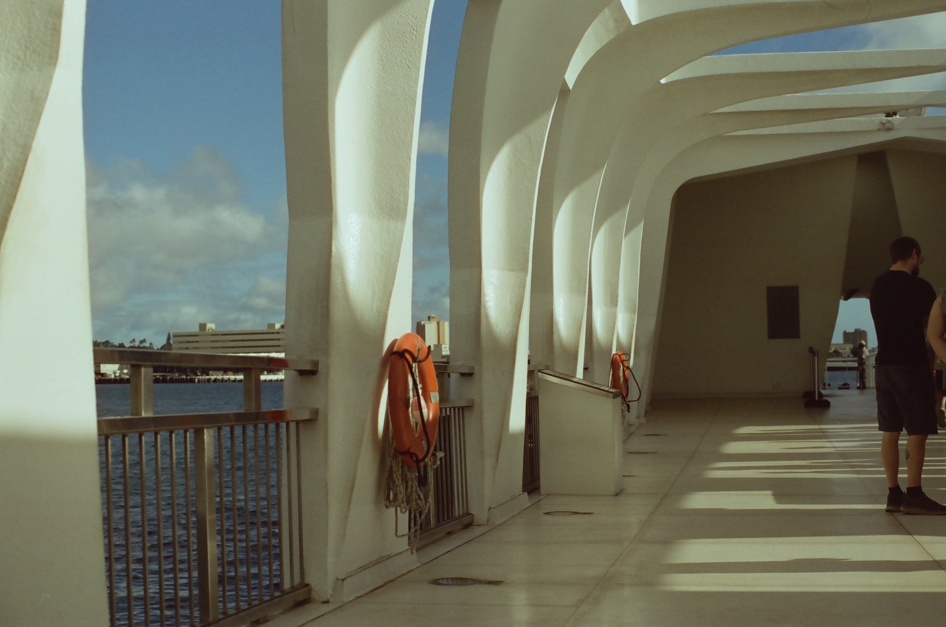 Repeating white arches cast long rectangular shadows along an open waterfront corridor beside blue water.