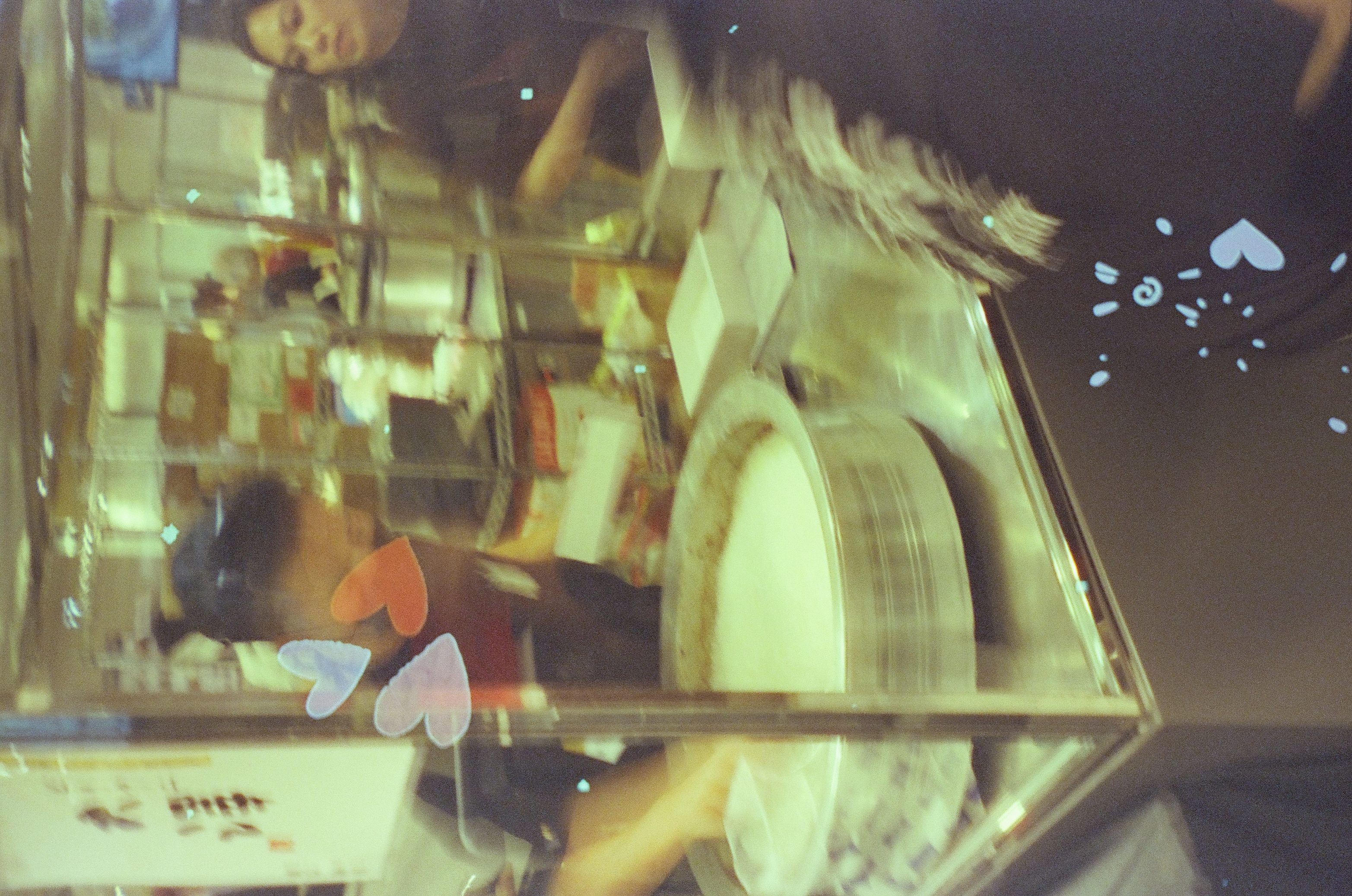 Through a glass display case, a person with hair in a bun bends over a wide pan of pale liquid, with small heart stickers on the case's metal frame.
