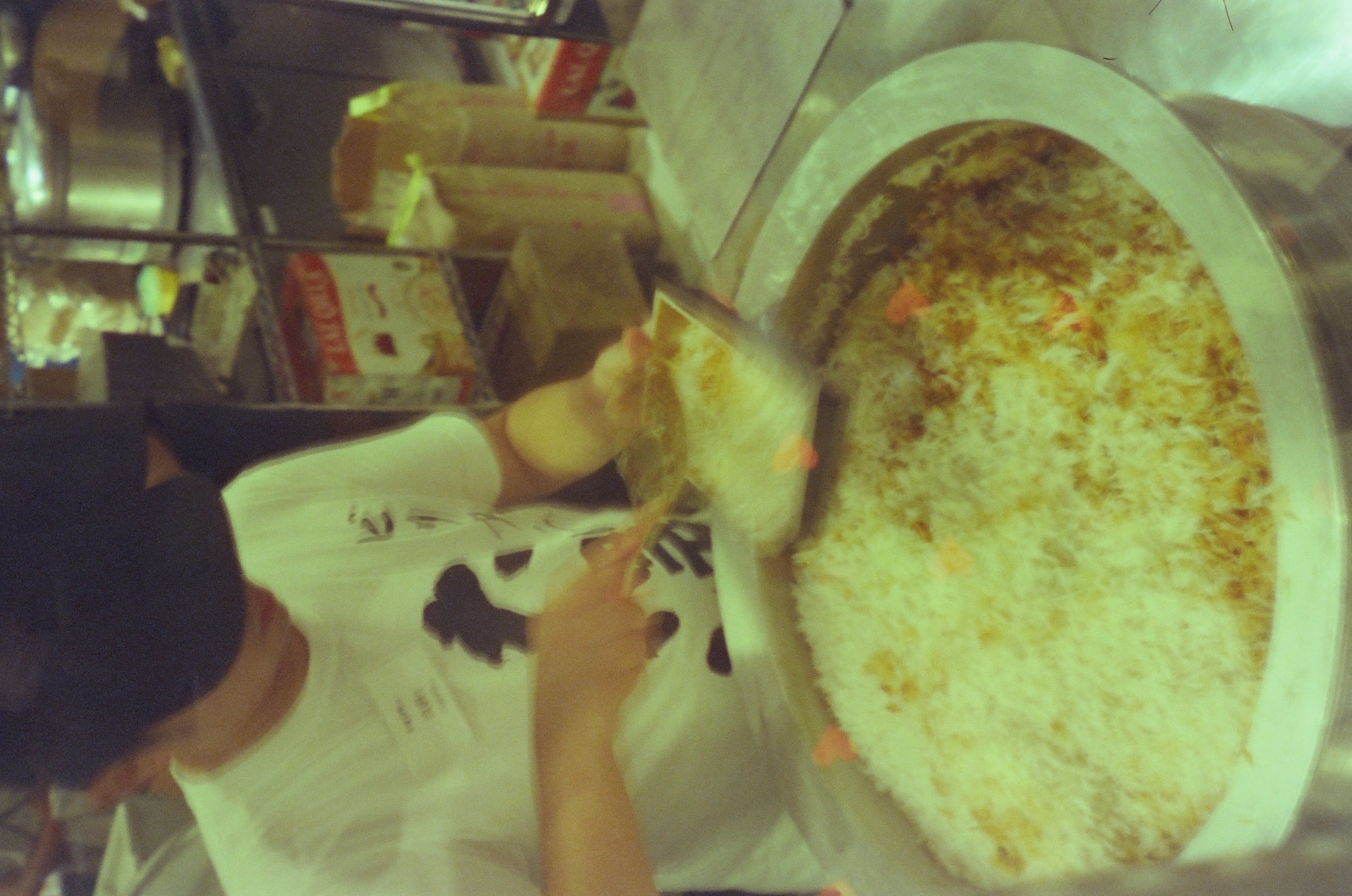 A cook in a white t-shirt and dark cap grates food above a large pan of pale shredded food, with one small orange heart-shaped mark at lower left.