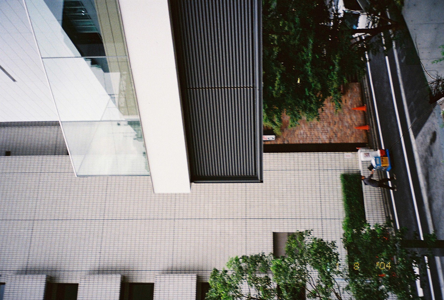 Seen from above, a worker pushes a blue-and-white cart along a street past orange traffic cones beneath a tiled building facade.