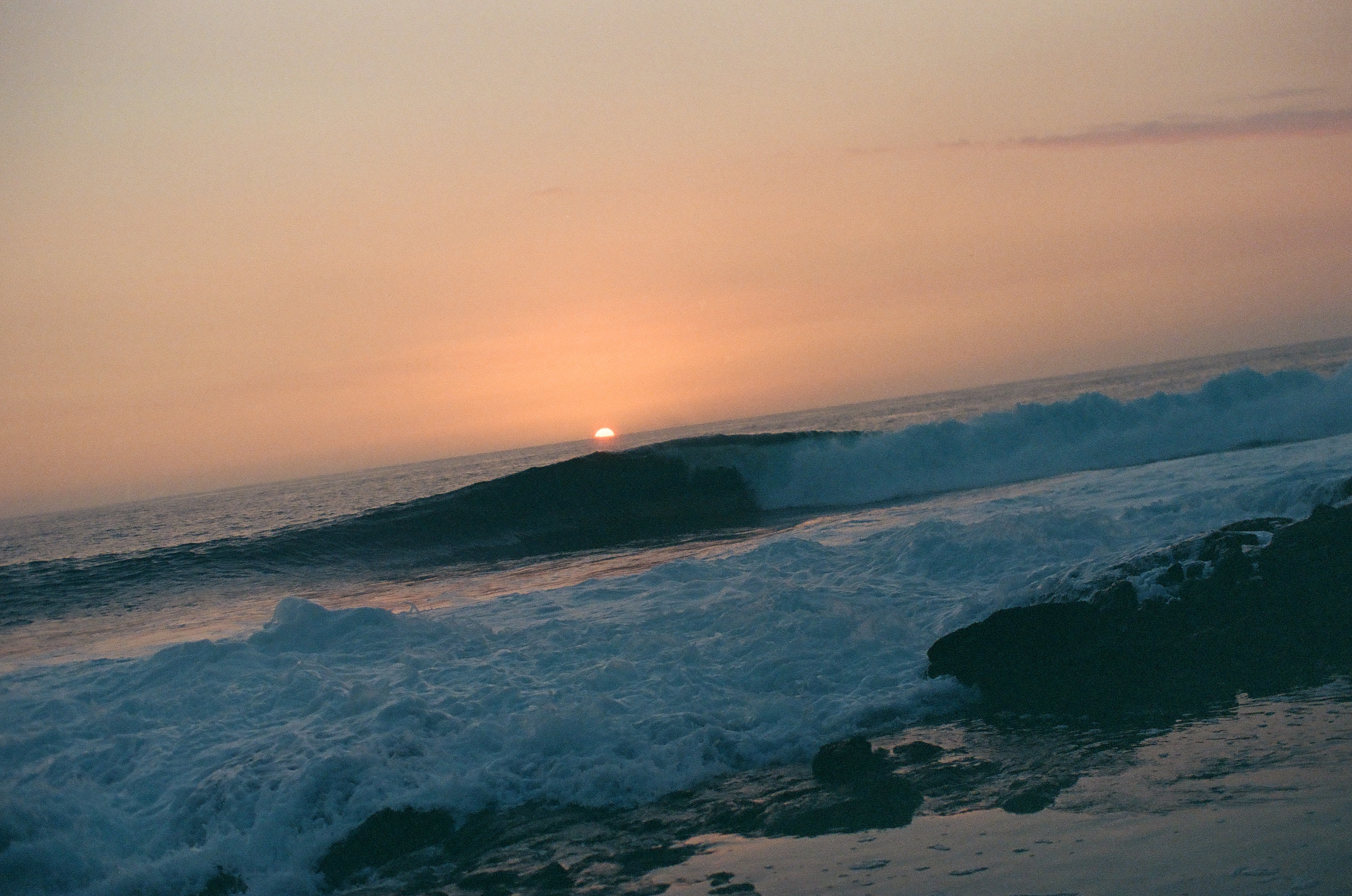 A dark blue wave lifts in front of a low orange sun above black shoreline rocks.