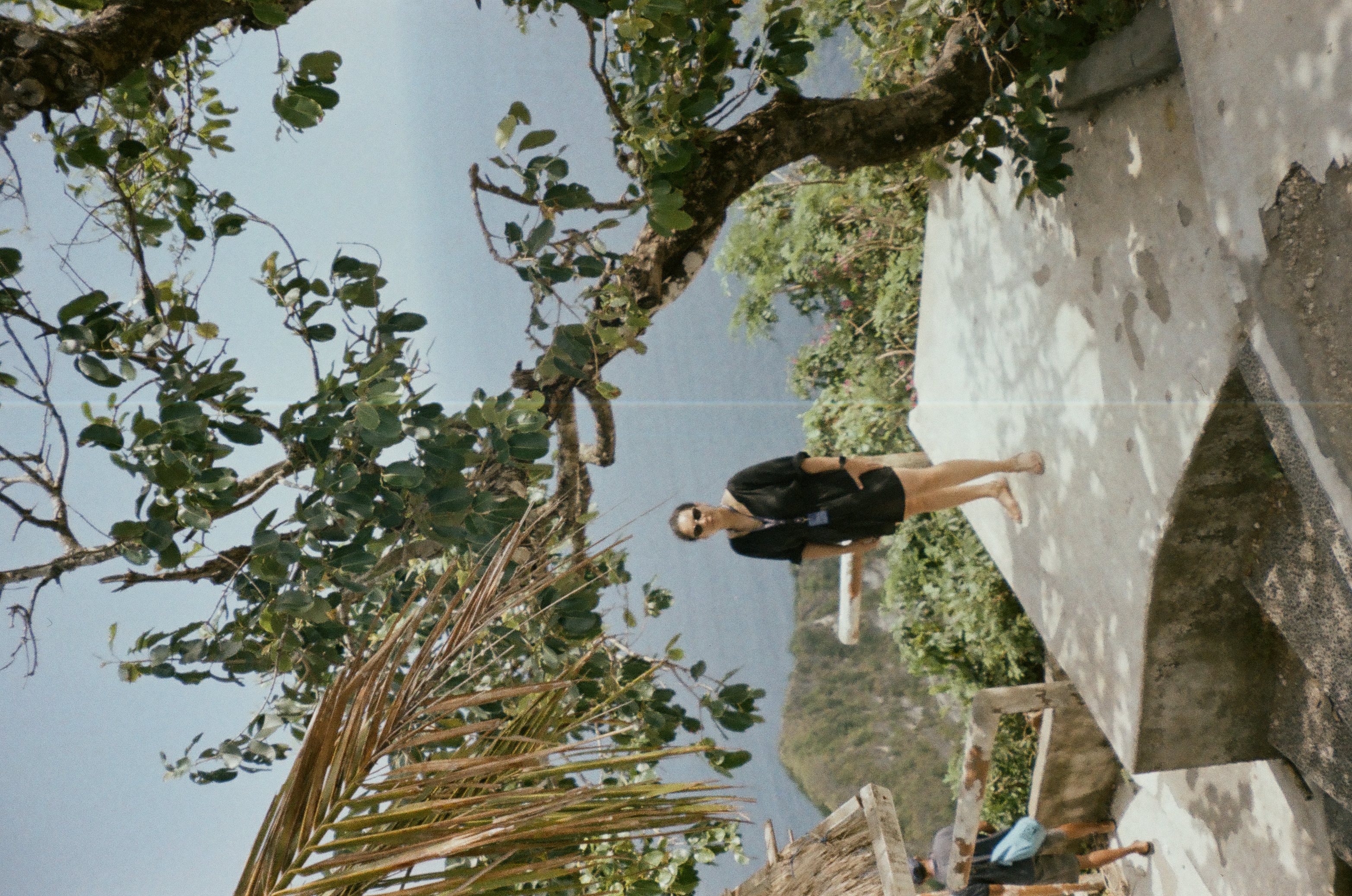 An adult in black stands on a concrete platform beneath spreading branches, with dense vegetation and hazy blue water beyond; another person is partly visible at lower left.