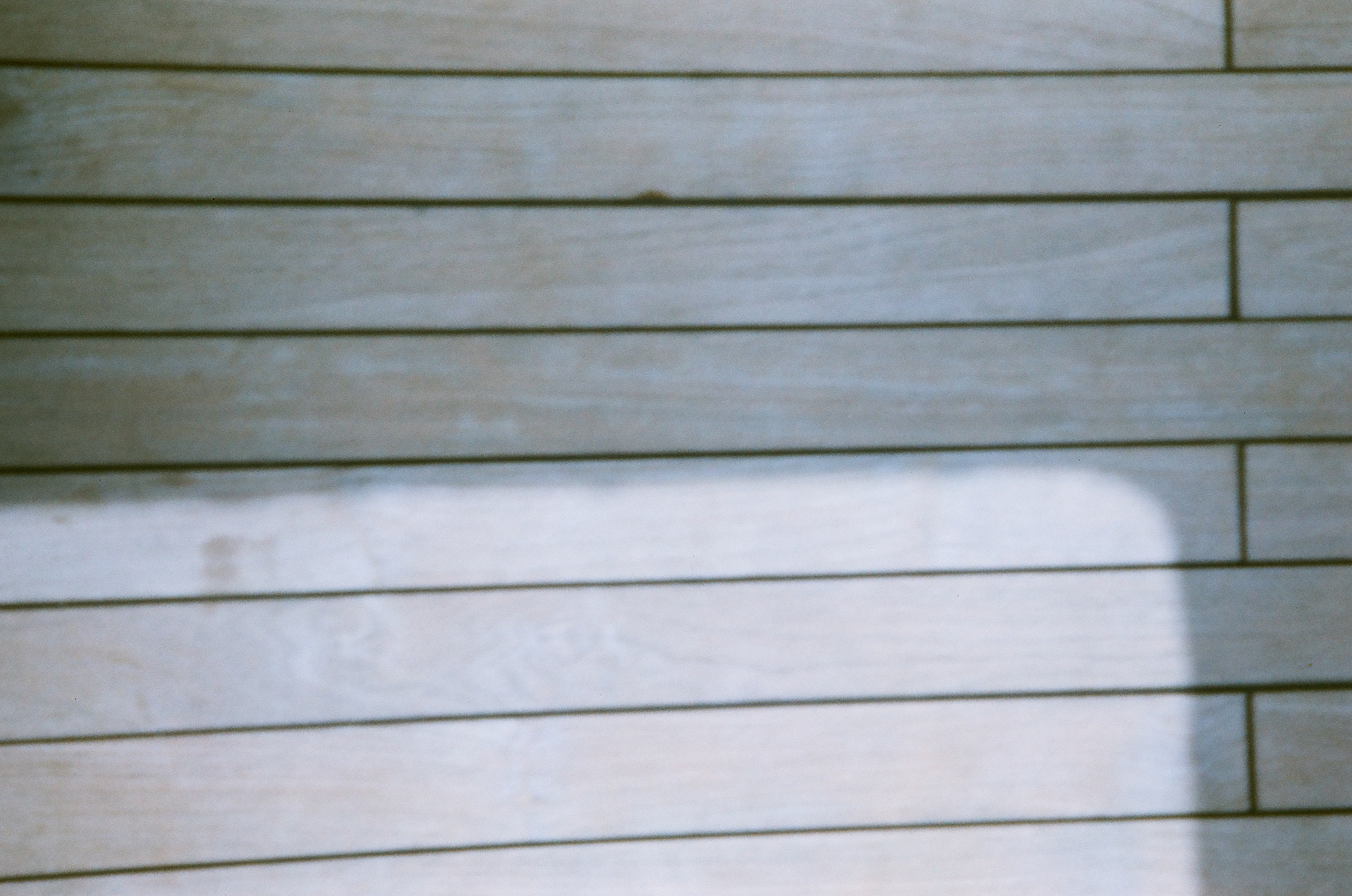 Parallel pale deck planks fill the frame as a brighter rectangular area and soft-edged shadow interrupt the lines.