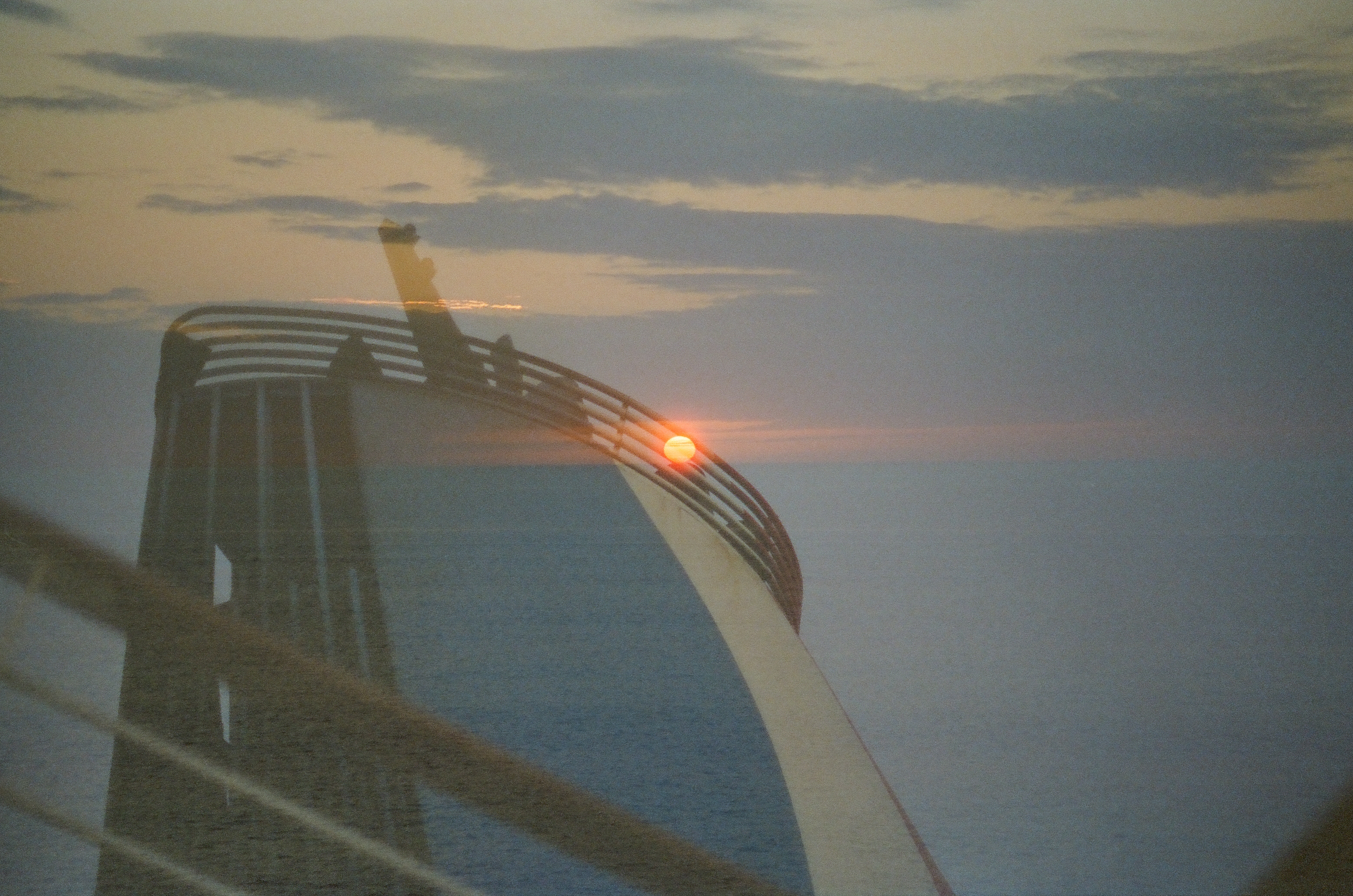 Translucent curved railings on a broad pale curving surface overlap a seascape where a small orange sun sits at the horizon by the rail.