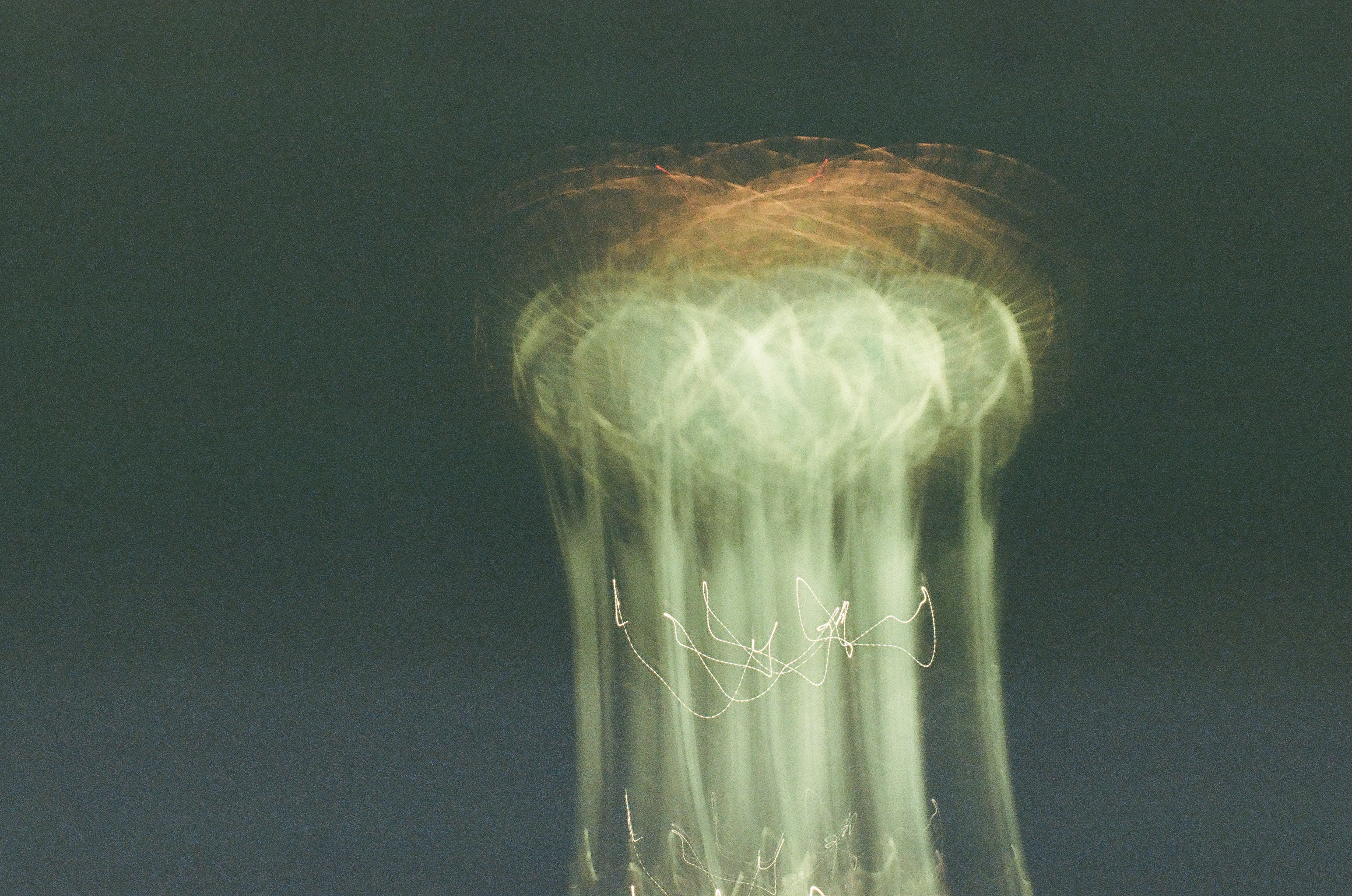 A pale tower smears upward into overlapping green-white rings while thin looping light trails cross its lower half.