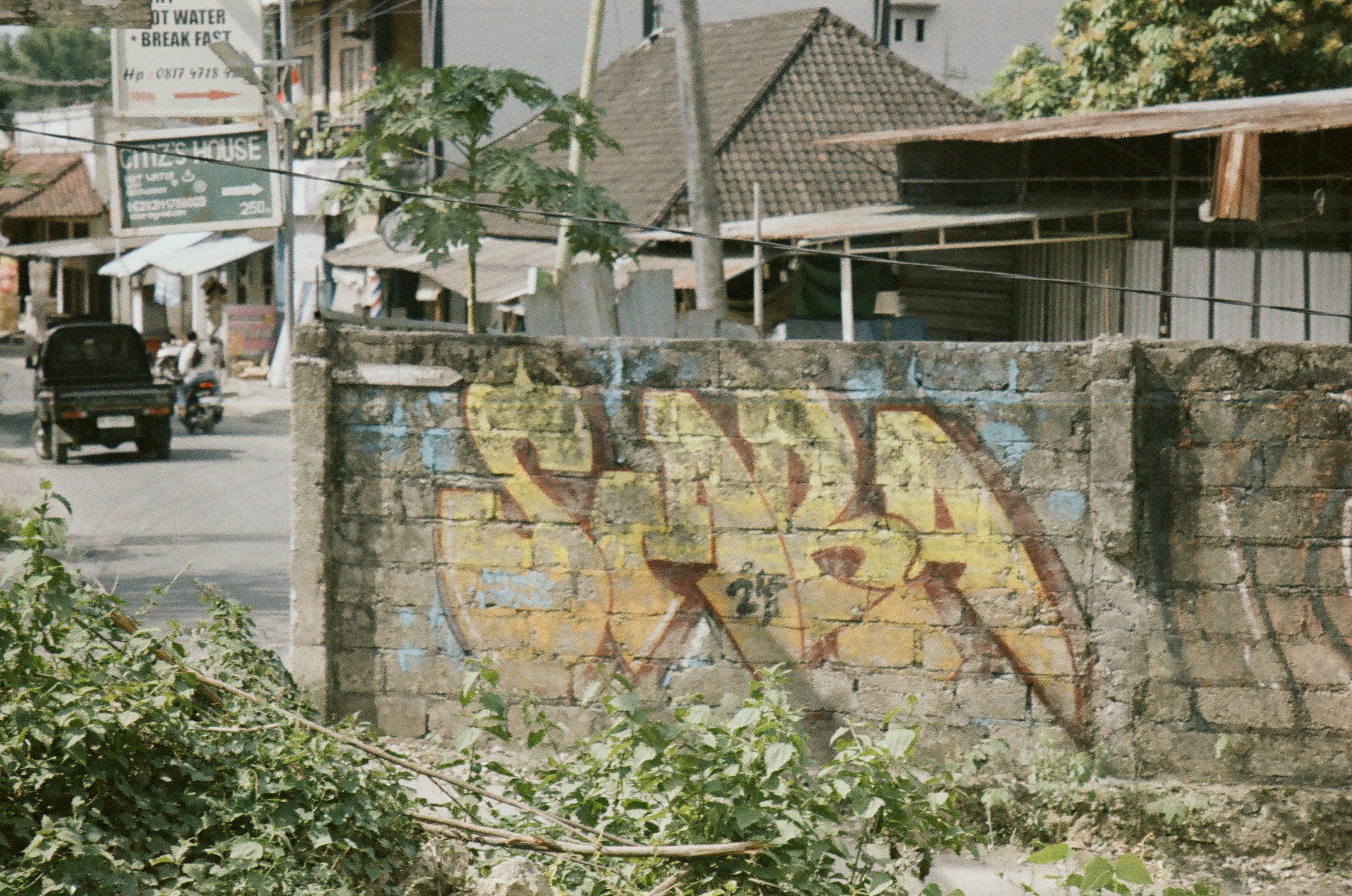 A graffitied concrete block wall borders a roadside lined with tiled roofs, leafy trees, signs, and vehicles.