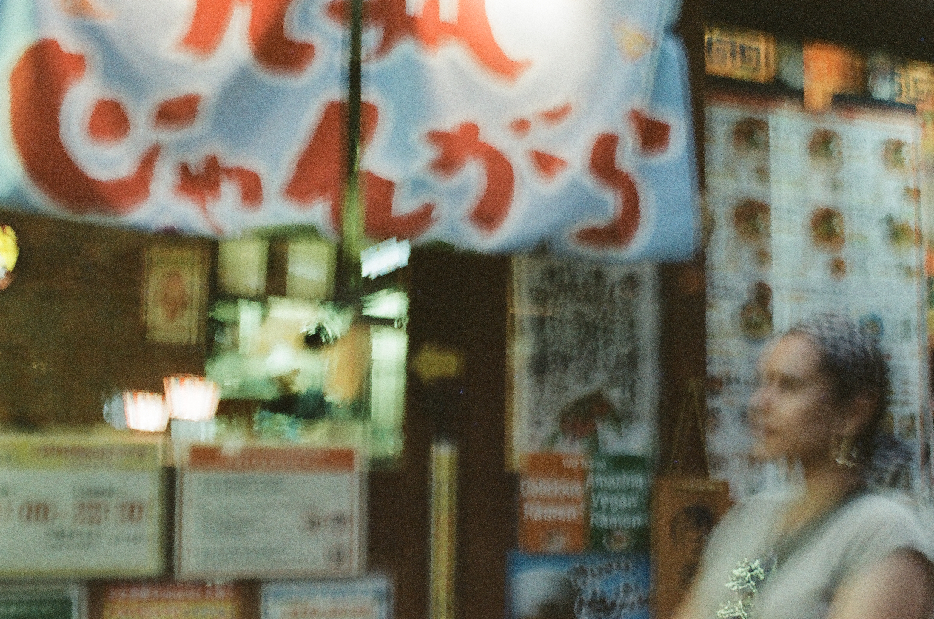 A white banner with red Japanese lettering hangs above a wall papered with menus reading 'Delicious Ramen' and 'Amazing Vegan Ramen.'