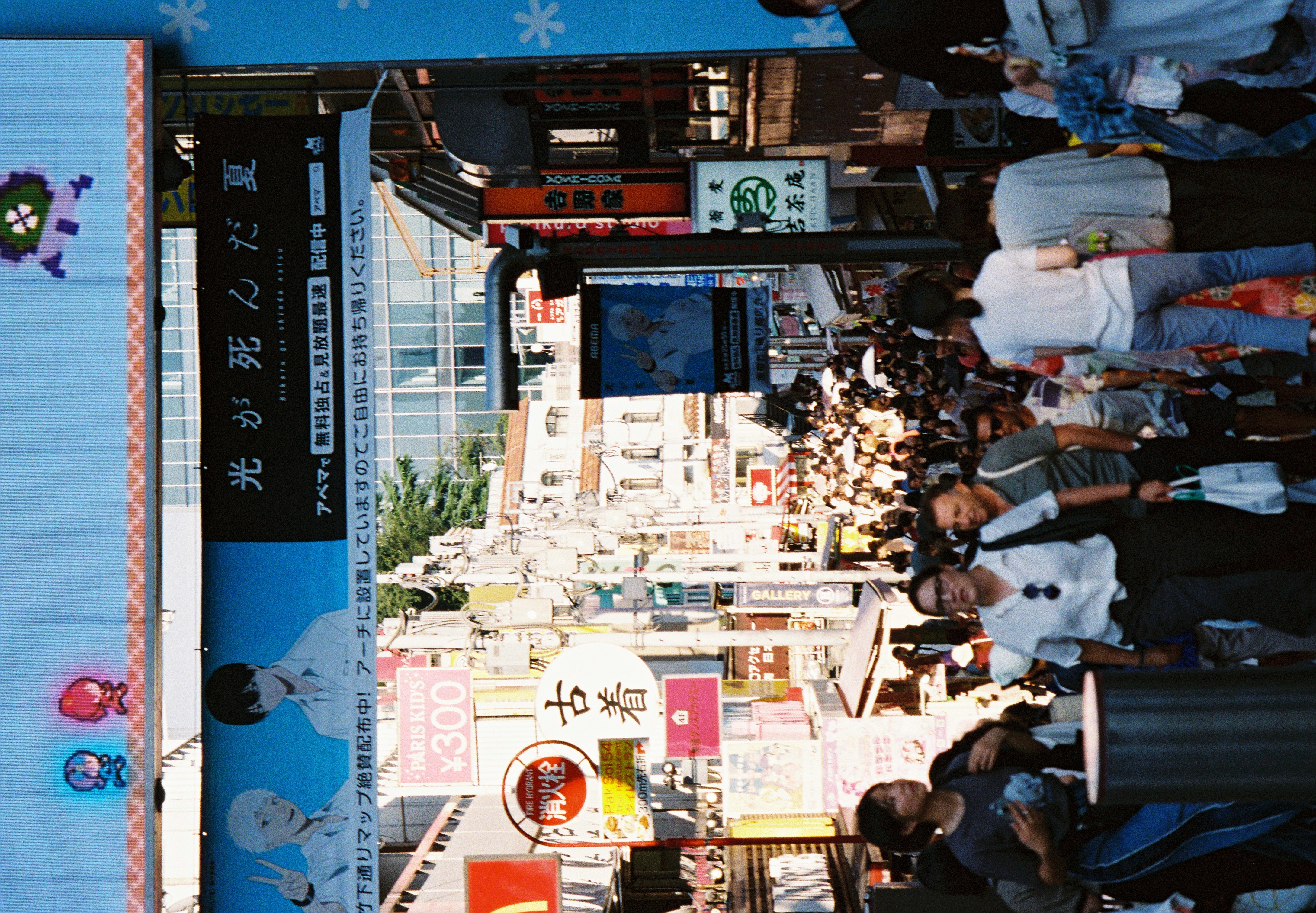 A dense crowd fills a narrow shopping street lined with stacked signage and an illustrated billboard, viewed from an elevated vantage near its entrance.