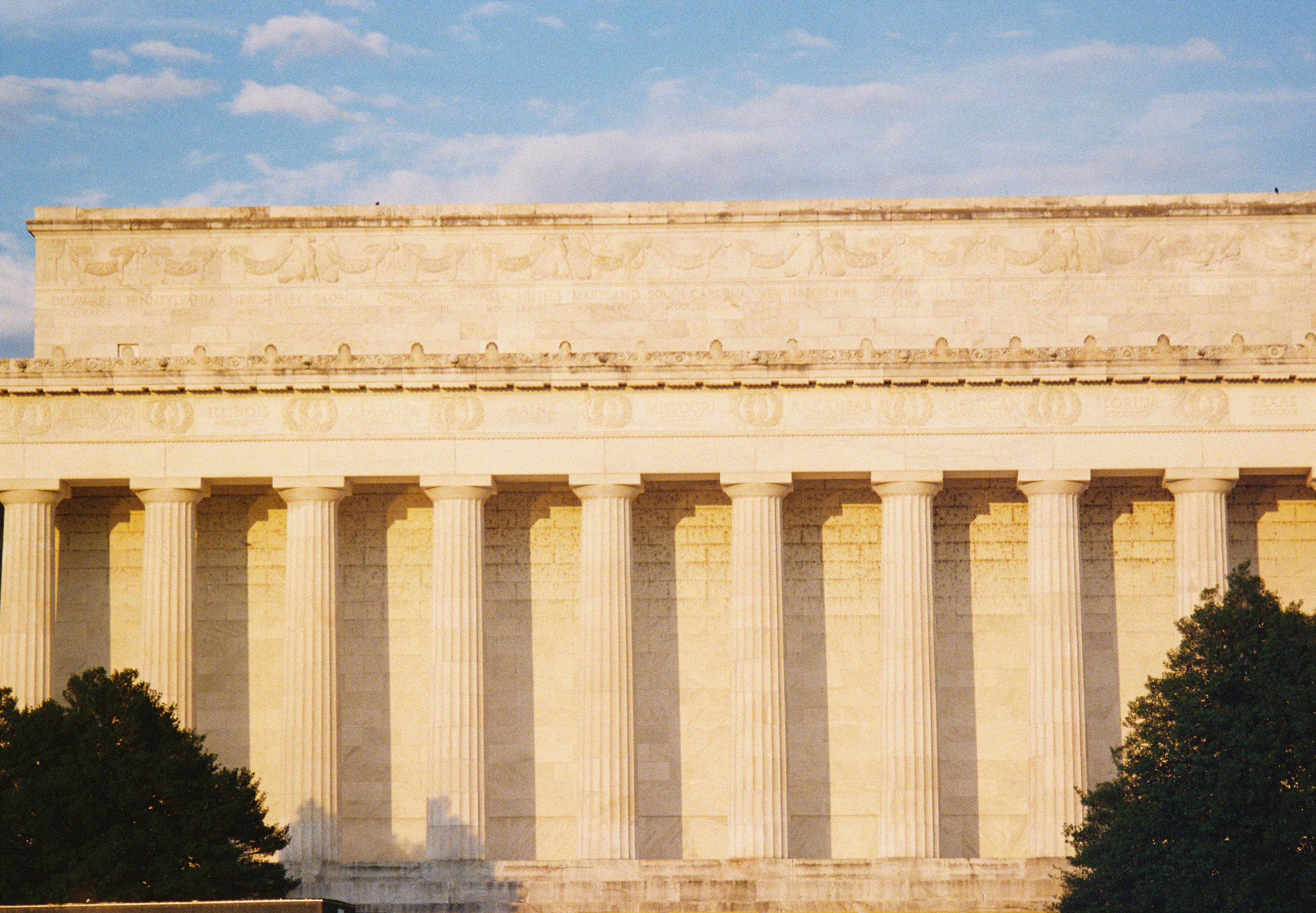 Sunlit colonnade of fluted columns beneath two carved bands of names including ILLINOIS, MAINE, MISSOURI, and FLORIDA, with dark treetops at the lower corners.