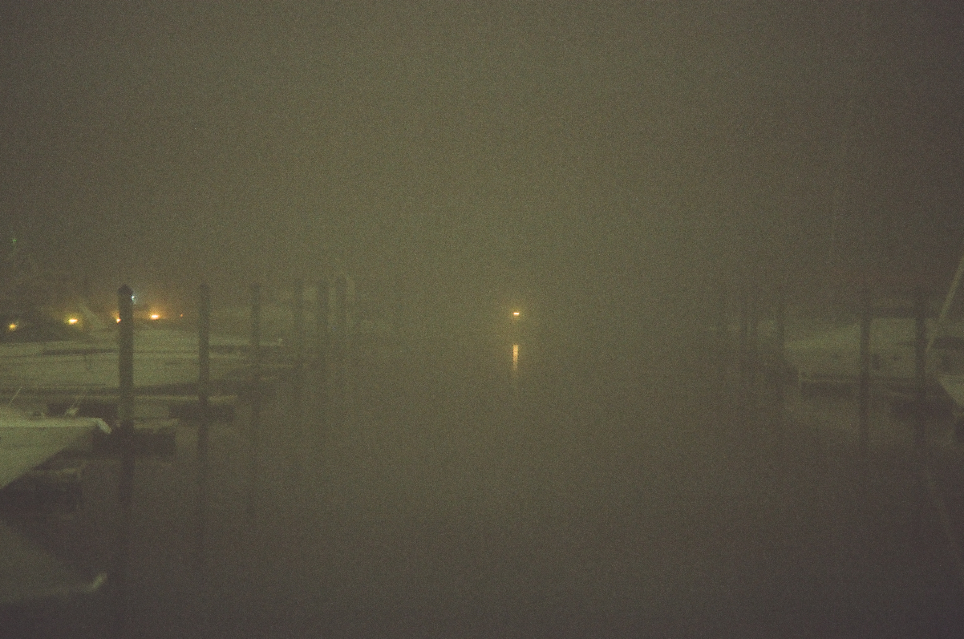 Dense fog nearly erases a marina at night, leaving only faint boat shapes at left and one distant light reflected in the water.
