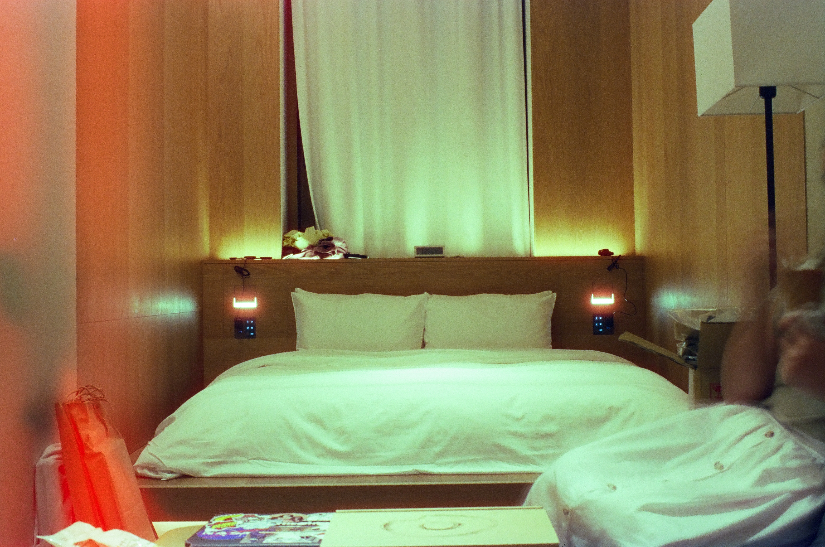 A white double bed with two pillows sits between wood-paneled walls under a backlit curtain, with a paper bag in the foreground and an orange glow at left.