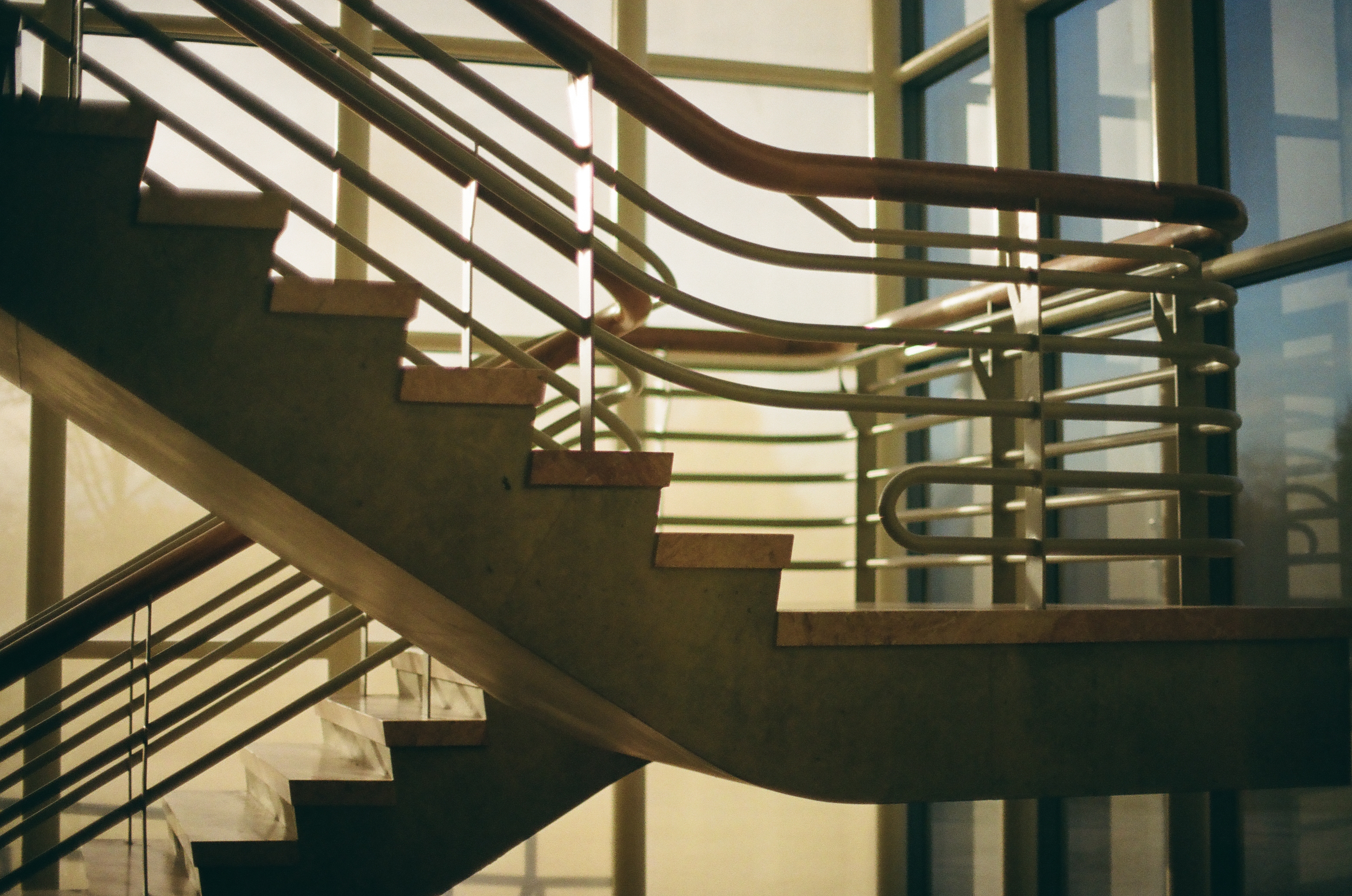 Concrete stair flights and rounded tubular rails overlap before tall gridded windows glowing with warm light.