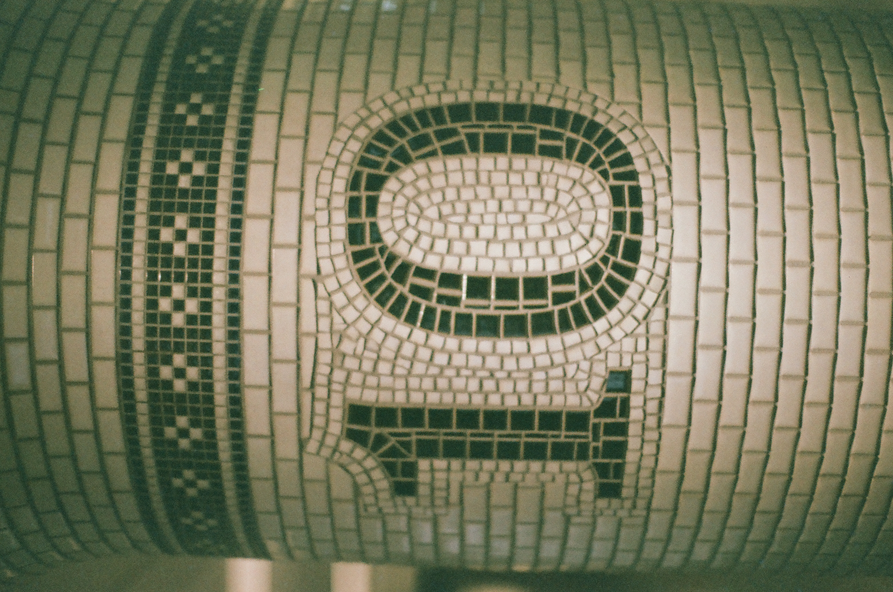 A stylized mosaic glyph in dark green tile fills a rounded column faced with pale tesserae and patterned banding.