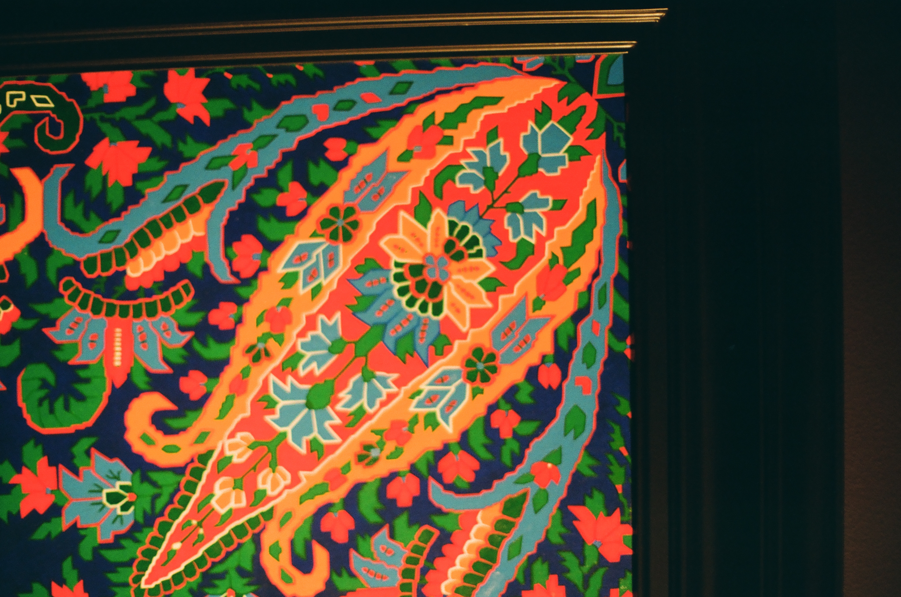 A large orange paisley shape filled with small blue and green flowers sits on a dark blue patterned ground beside a dark frame edge.