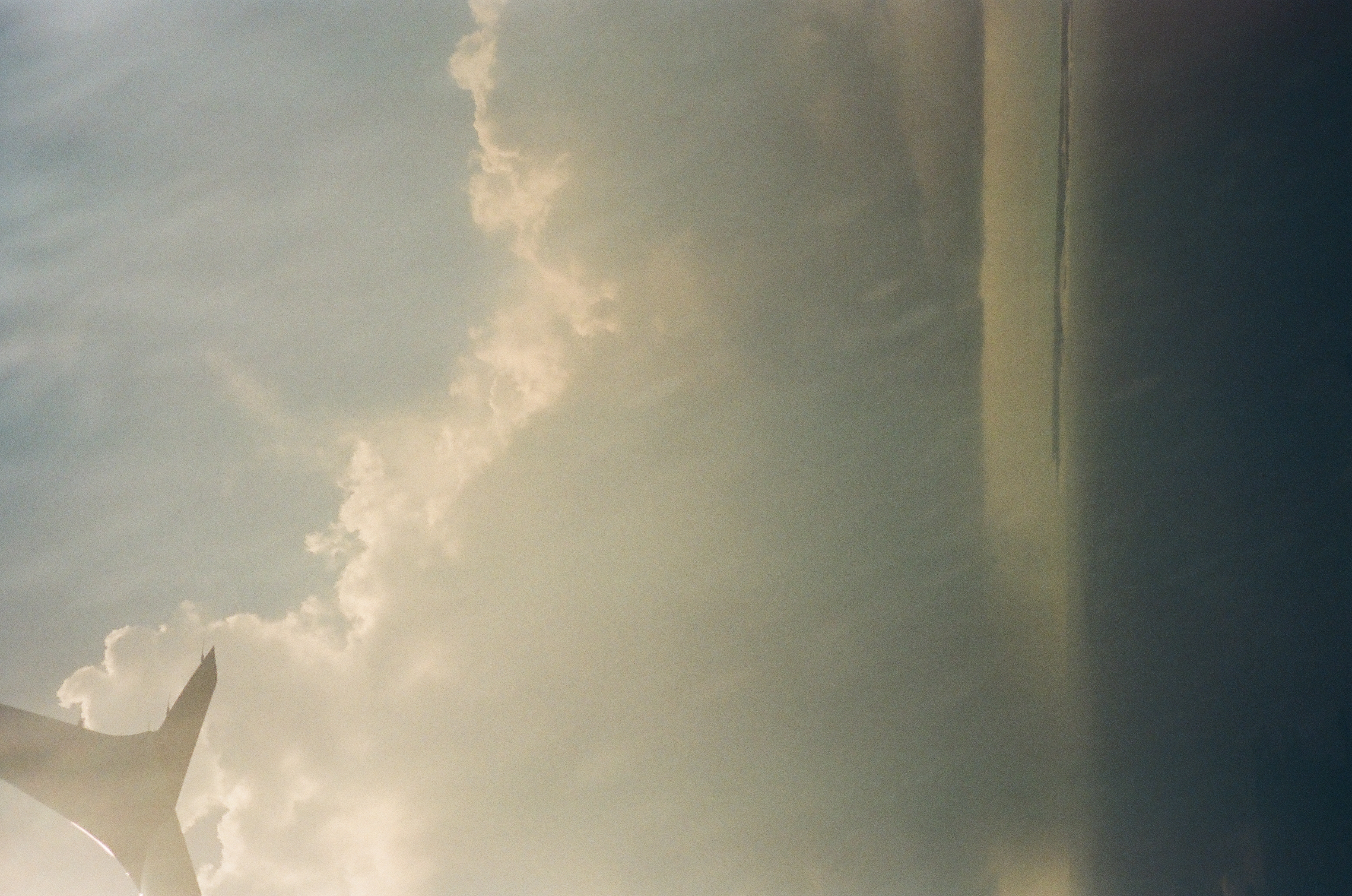 An aircraft wing enters at upper left above large clouds, vertical gray bands, a thin horizon, and dark water.