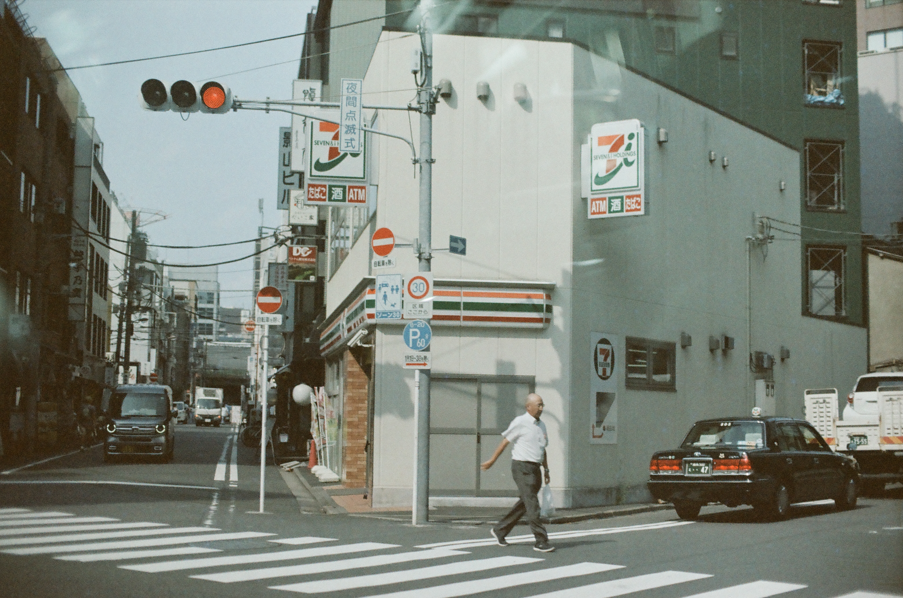 A man in a white shirt crosses a striped crosswalk beside a convenience store with green, orange, and red signage, as a dark car waits at the curb.